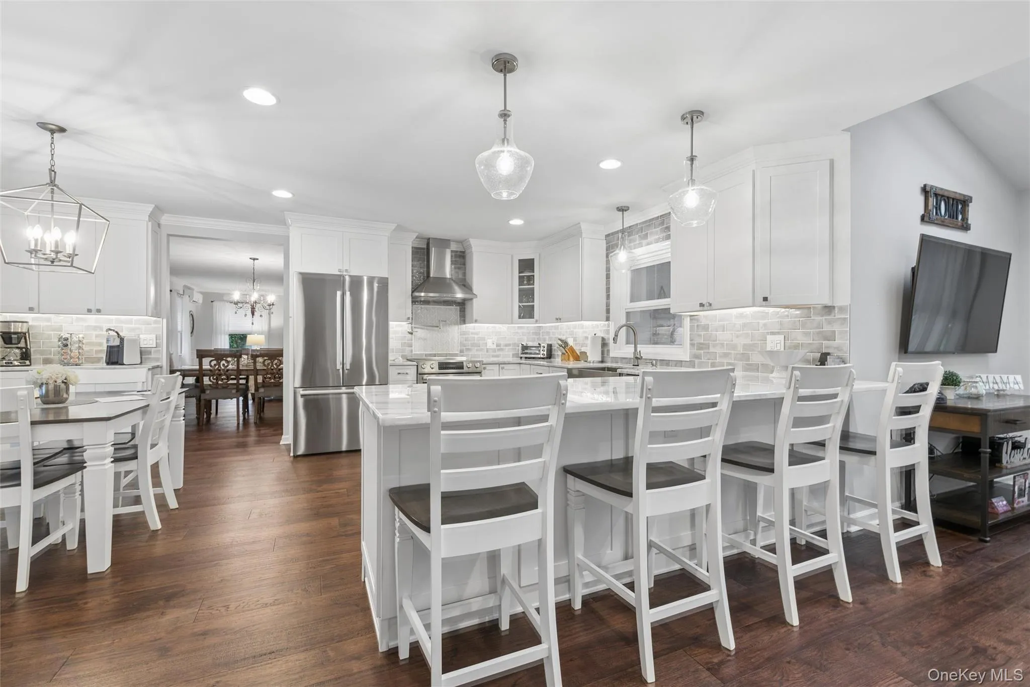 Bright kitchen featuring white cabinetry, a large center island with seating, stainless steel appliances, a tile backsplash, and wood-finish flooring Bright kitchen featuring white cabinetry, a large center island with seating, stainless steel appliances, a tile backsplash, and wood-finish flooring
