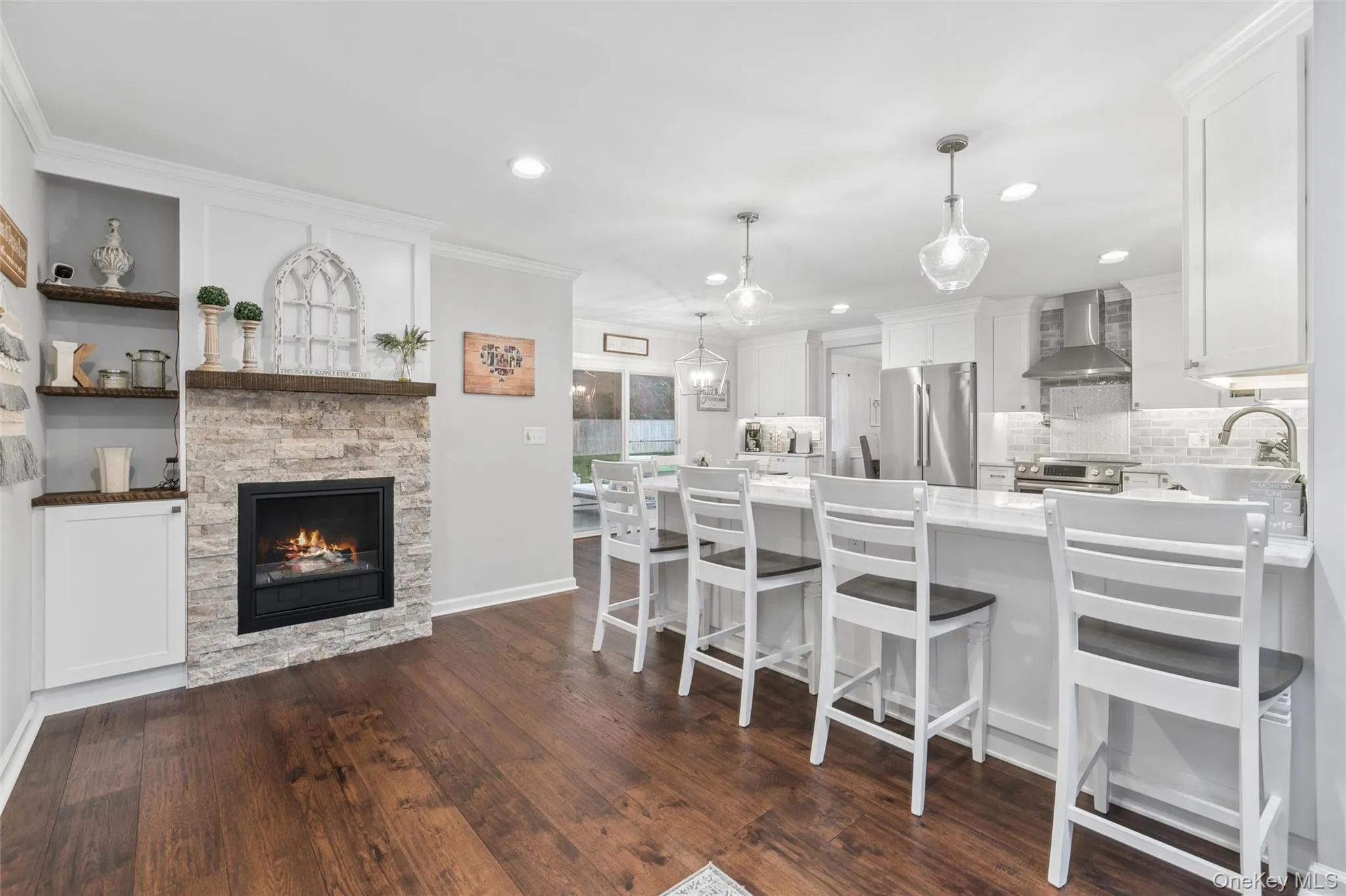 Spacious interior featuring wood-finish flooring, a stone-front fireplace with wood mantle, built-in shelving, a kitchen island with seating, and pendant lighting Spacious interior featuring wood-finish flooring, a stone-front fireplace with wood mantle, built-in shelving, a kitchen island with seating, and pendant lighting