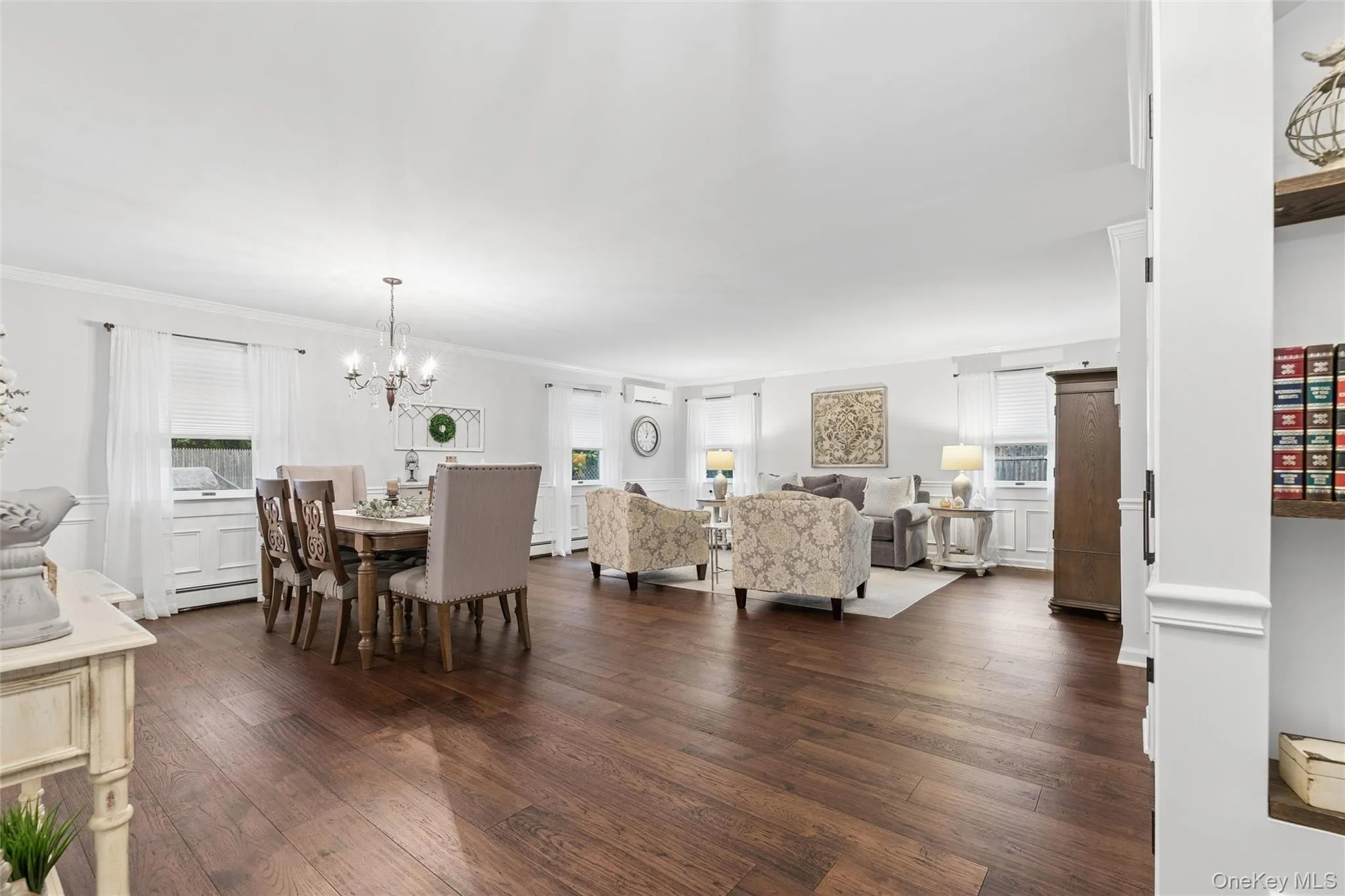 Open-concept living and dining area featuring wood-finish flooring, white paneled walls, crown molding, and multiple windows Open-concept living and dining area featuring wood-finish flooring, white paneled walls, crown molding, and multiple windows
