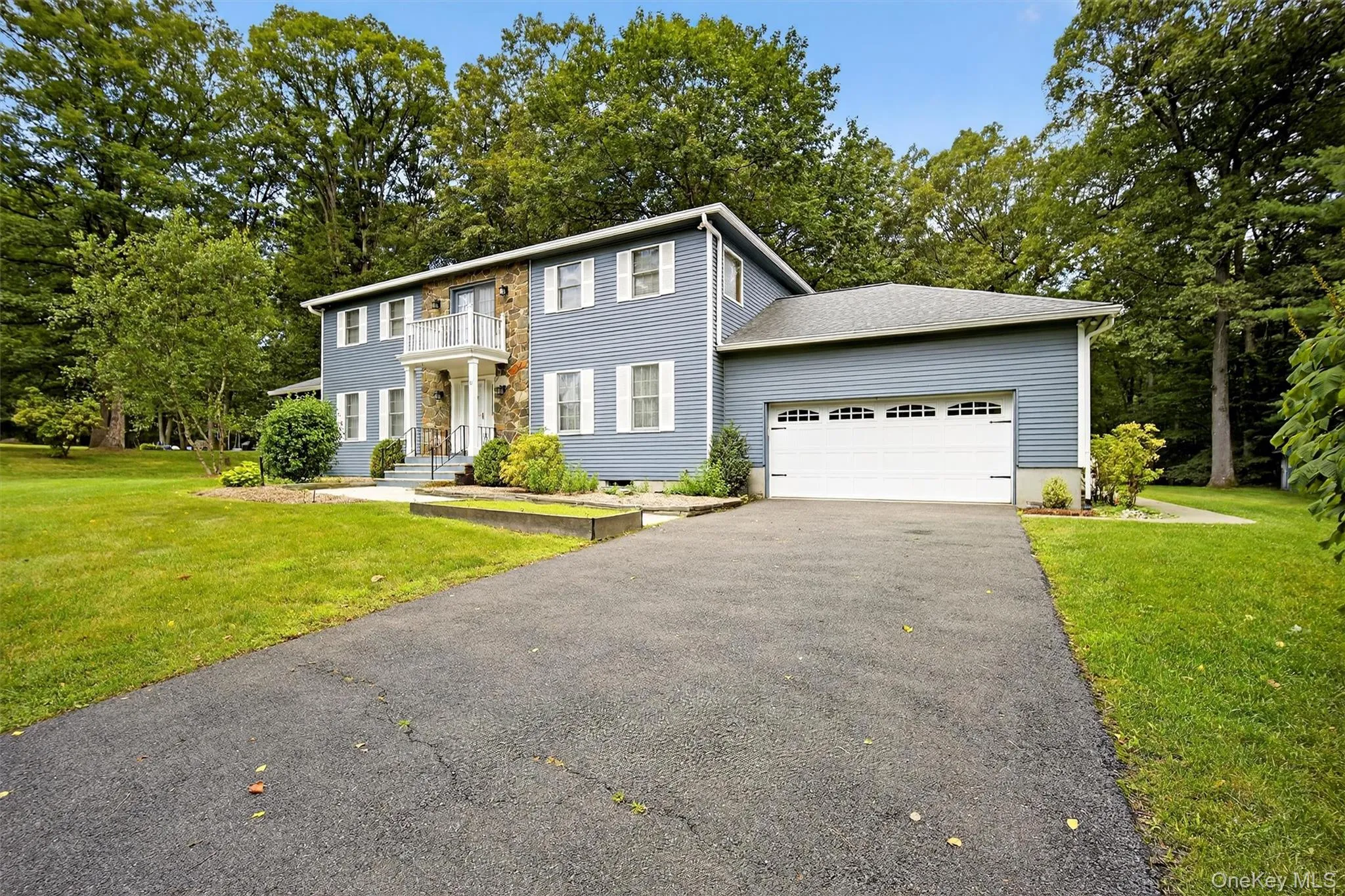 View of front facade with a balcony, a garage, asphalt driveway, a front lawn, and view of wooded area View of front facade with a balcony, a garage, asphalt driveway, a front lawn, and view of wooded area