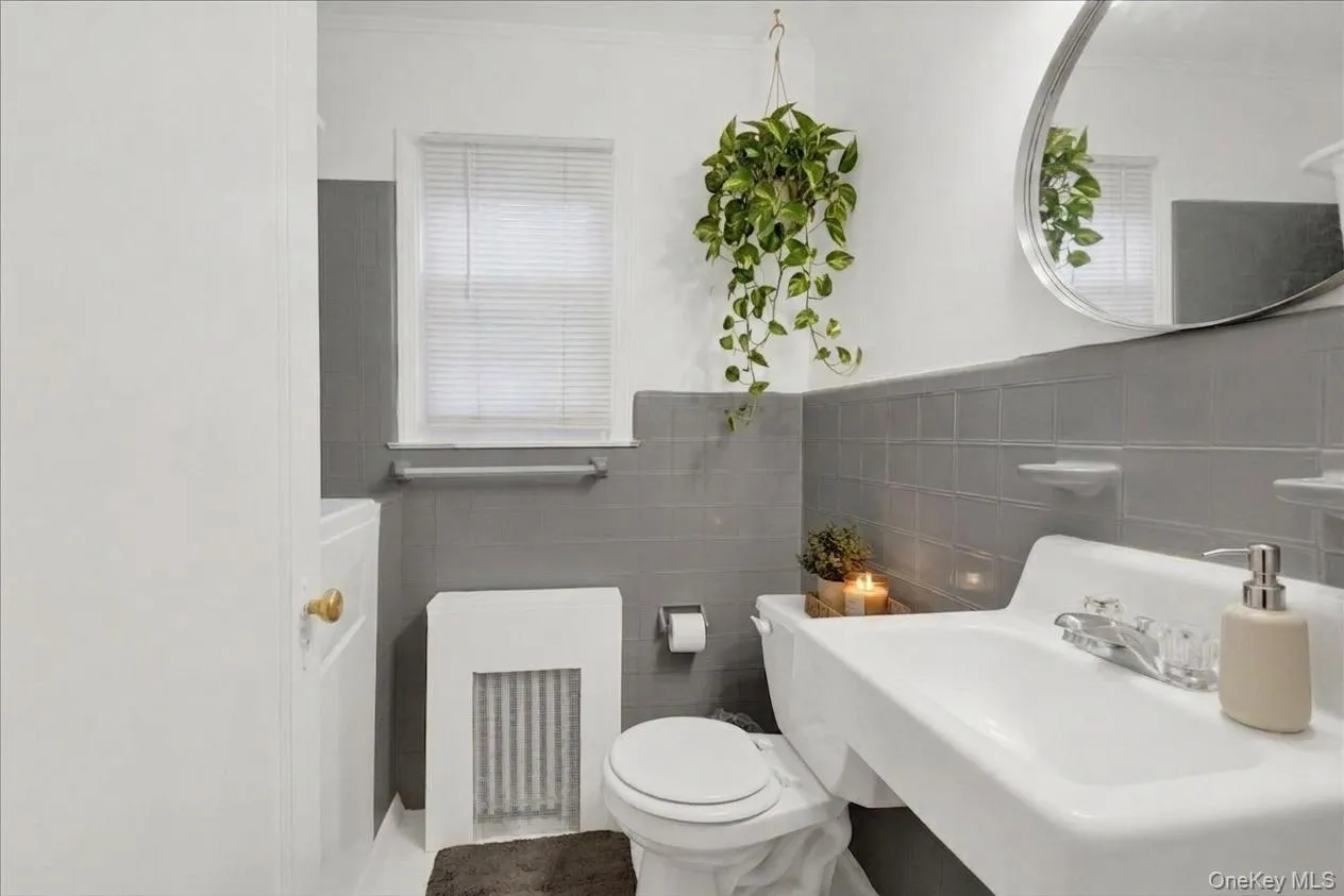 First floor full bathroom featuring half-tiled grey walls, a wall-mounted sink with clear acrylic faucets, and a rounded mirror First floor full bathroom featuring half-tiled grey walls, a wall-mounted sink with clear acrylic faucets, and a rounded mirror