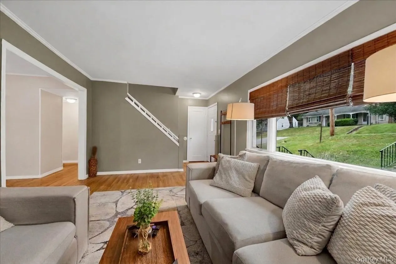 Spacious living area featuring expansive windows with bamboo shades, wood-finish flooring, and a visible staircase Spacious living area featuring expansive windows with bamboo shades, wood-finish flooring, and a visible staircase