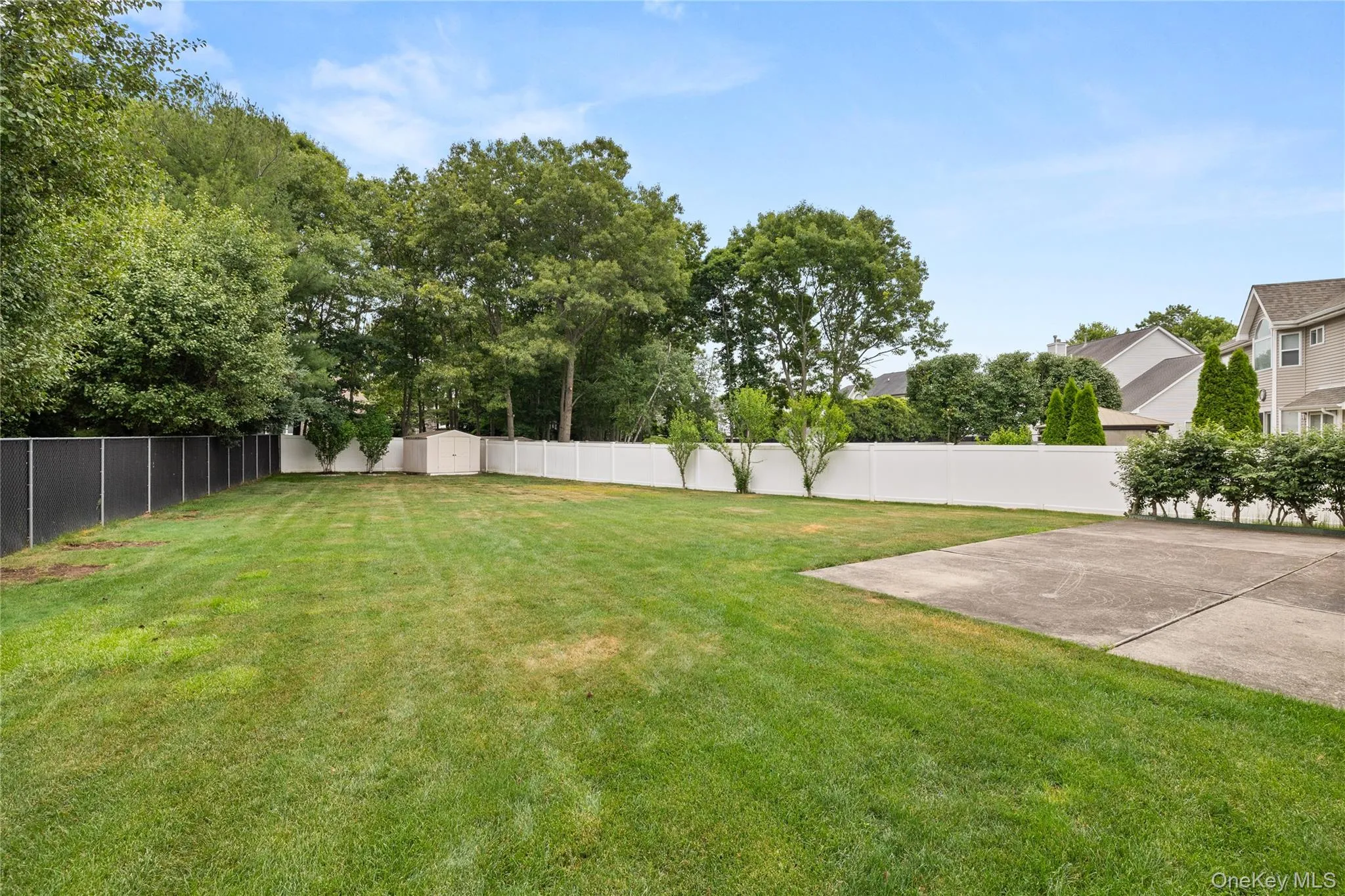 Expansive backyard featuring a large concrete patio, mature perimeter trees, and a combination of white privacy fencing and black chain-link fencing Expansive backyard featuring a large concrete patio, mature perimeter trees, and a combination of white privacy fencing and black chain-link fencing