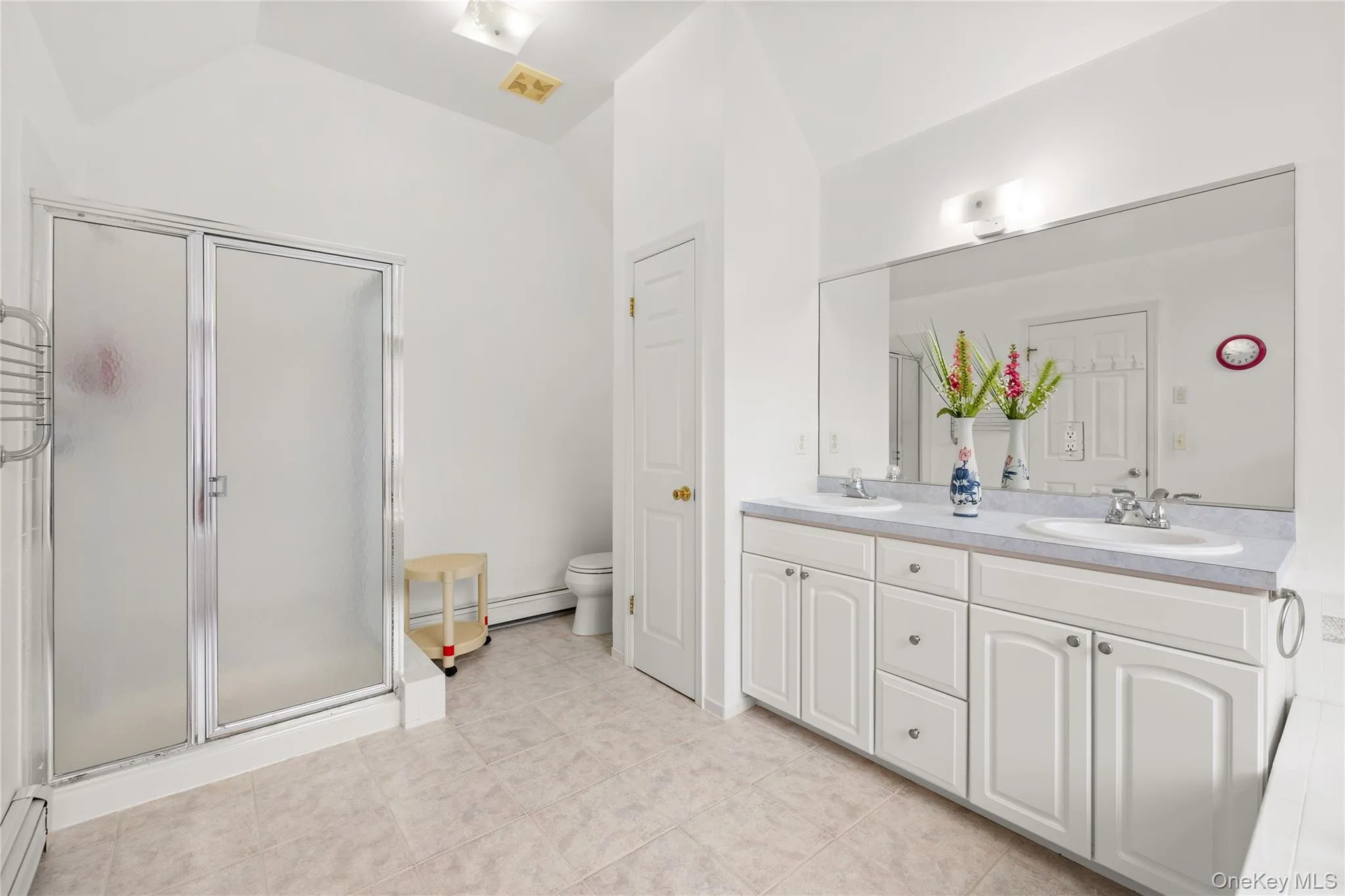 Primary bathroom featuring a dual vanity with white cabinetry and integrated sinks, a large wall-mounted mirror, a standing shower with frosted glass enclosure, and light-toned tile flooring Primary bathroom featuring a dual vanity with white cabinetry and integrated sinks, a large wall-mounted mirror, a standing shower with frosted glass enclosure, and light-toned tile flooring