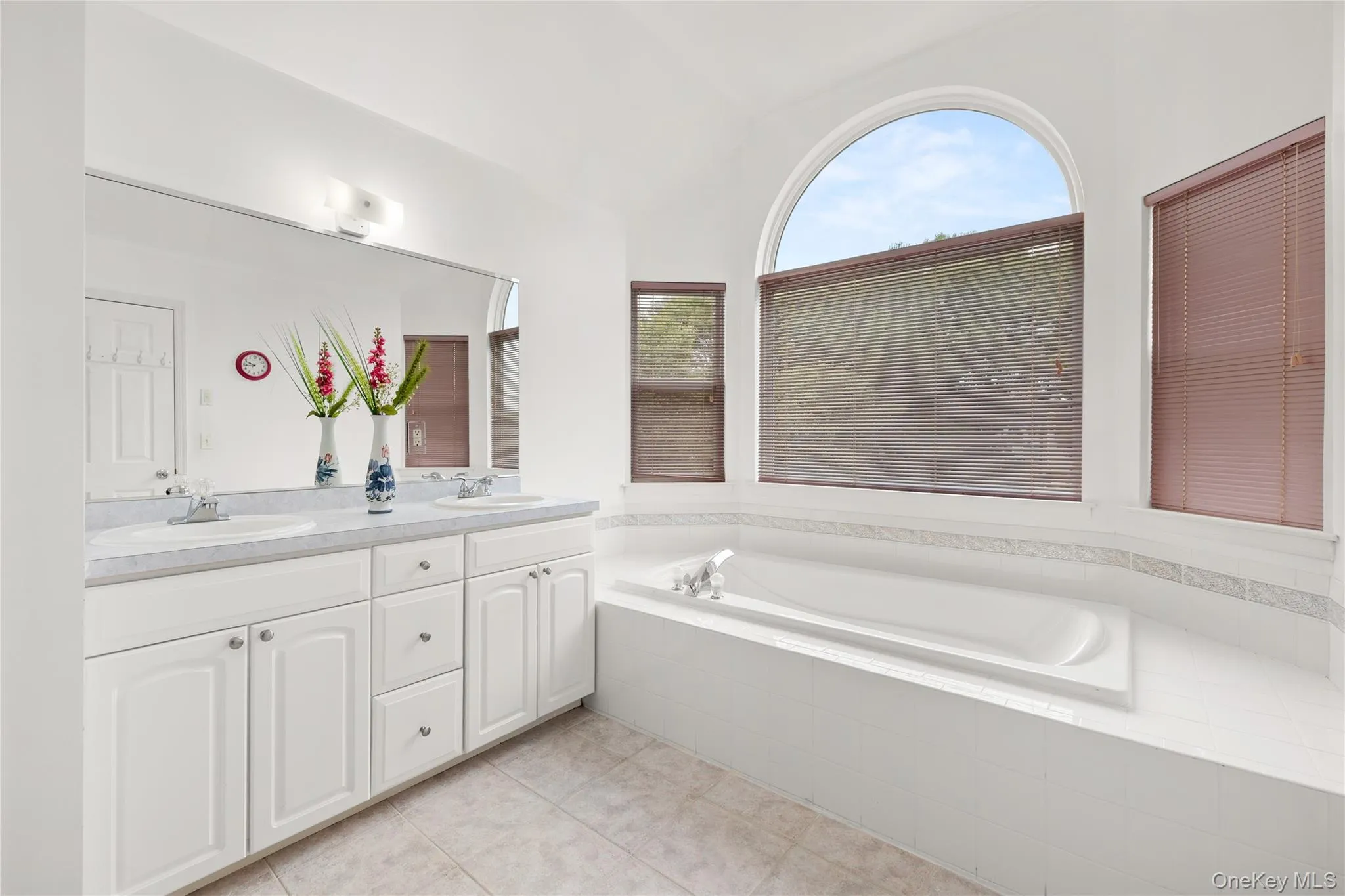 Primary bathroom featuring a built-in soaking tub, a large arched window, a double vanity with ample storage, and tile flooring Primary bathroom featuring a built-in soaking tub, a large arched window, a double vanity with ample storage, and tile flooring