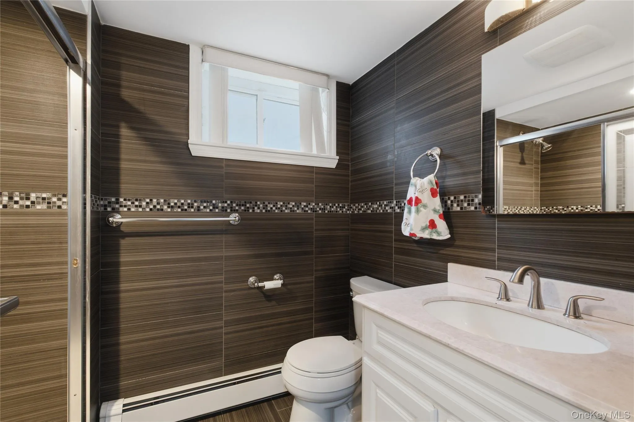 Bathroom featuring dark wood-finish tile walls with a mosaic accent band, white vanity with integrated sink, brushed nickel fixtures, and a glass-enclosed shower Bathroom featuring dark wood-finish tile walls with a mosaic accent band, white vanity with integrated sink, brushed nickel fixtures, and a glass-enclosed shower