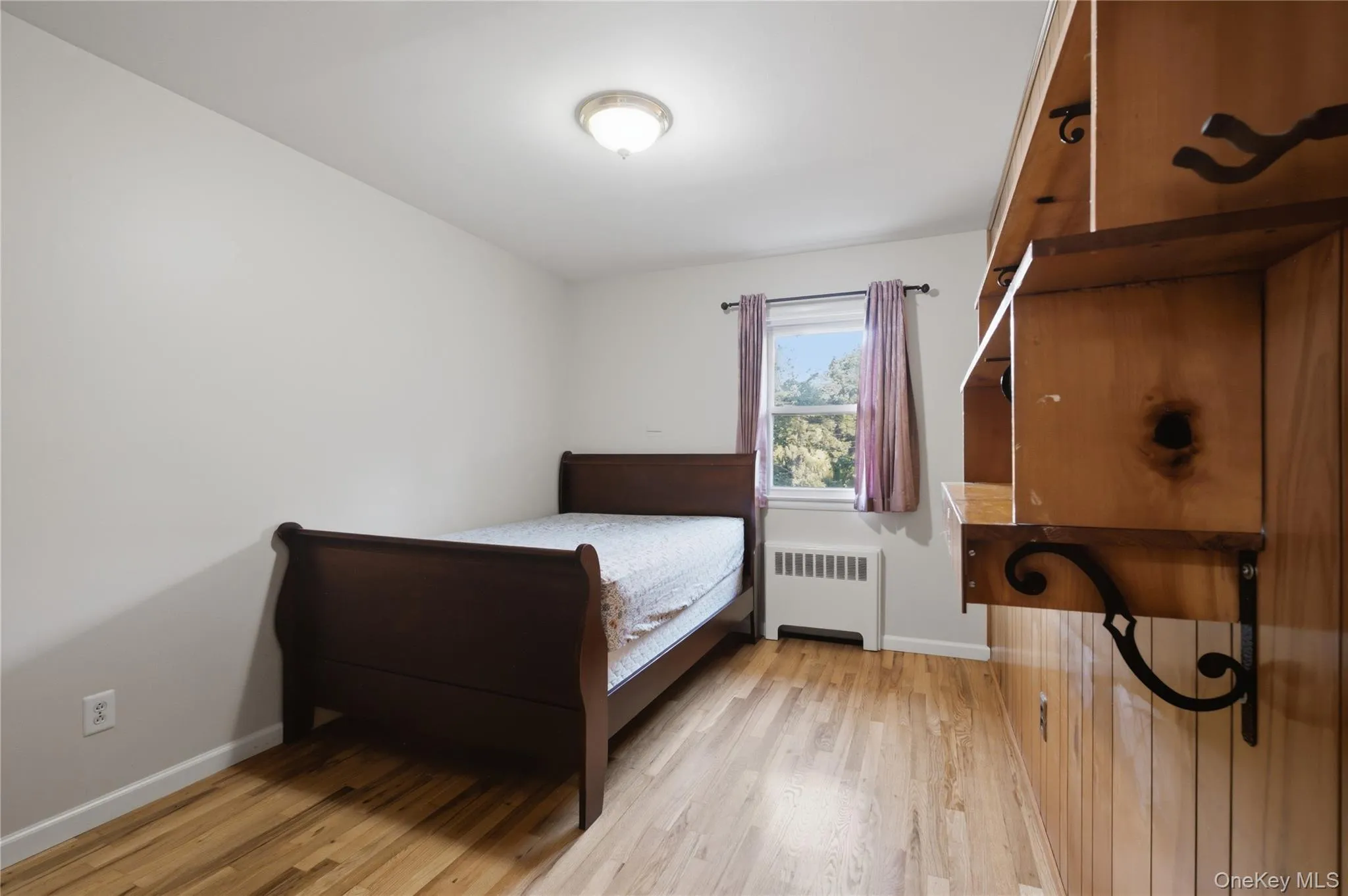 Room featuring wood-finish flooring, a double-hung window, a built-in wall unit with shelving, and a ceiling-mounted light fixture Room featuring wood-finish flooring, a double-hung window, a built-in wall unit with shelving, and a ceiling-mounted light fixture