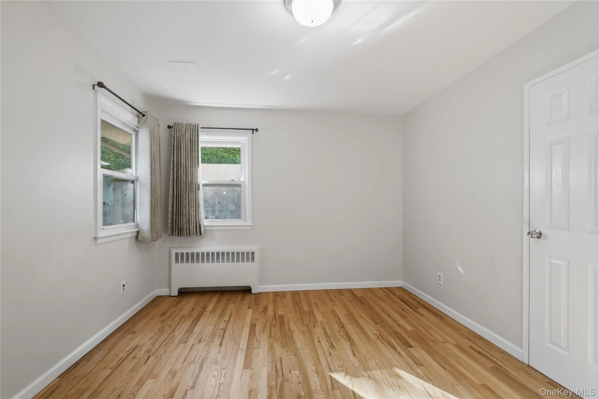 Room featuring wood flooring, neutral wall tones, two windows with white trim, a baseboard heating unit, and a flush-mount ceiling light Room featuring wood flooring, neutral wall tones, two windows with white trim, a baseboard heating unit, and a flush-mount ceiling light