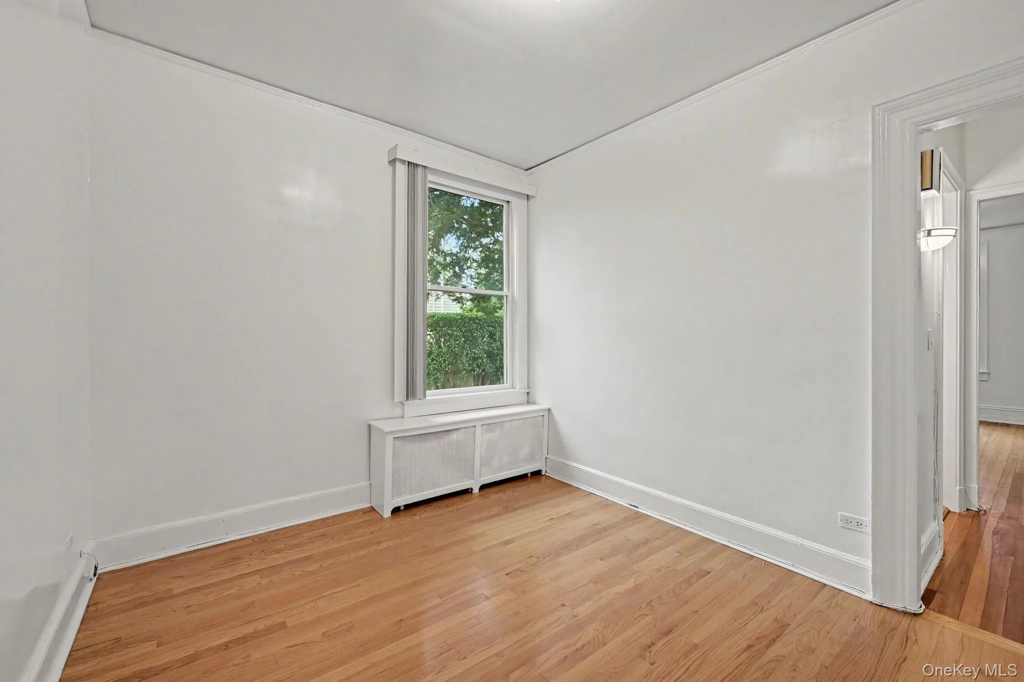 Bright bedroom featuring a window with exterior foliage, wood-finish flooring, white baseboards, and a radiator cover Bright bedroom featuring a window with exterior foliage, wood-finish flooring, white baseboards, and a radiator cover