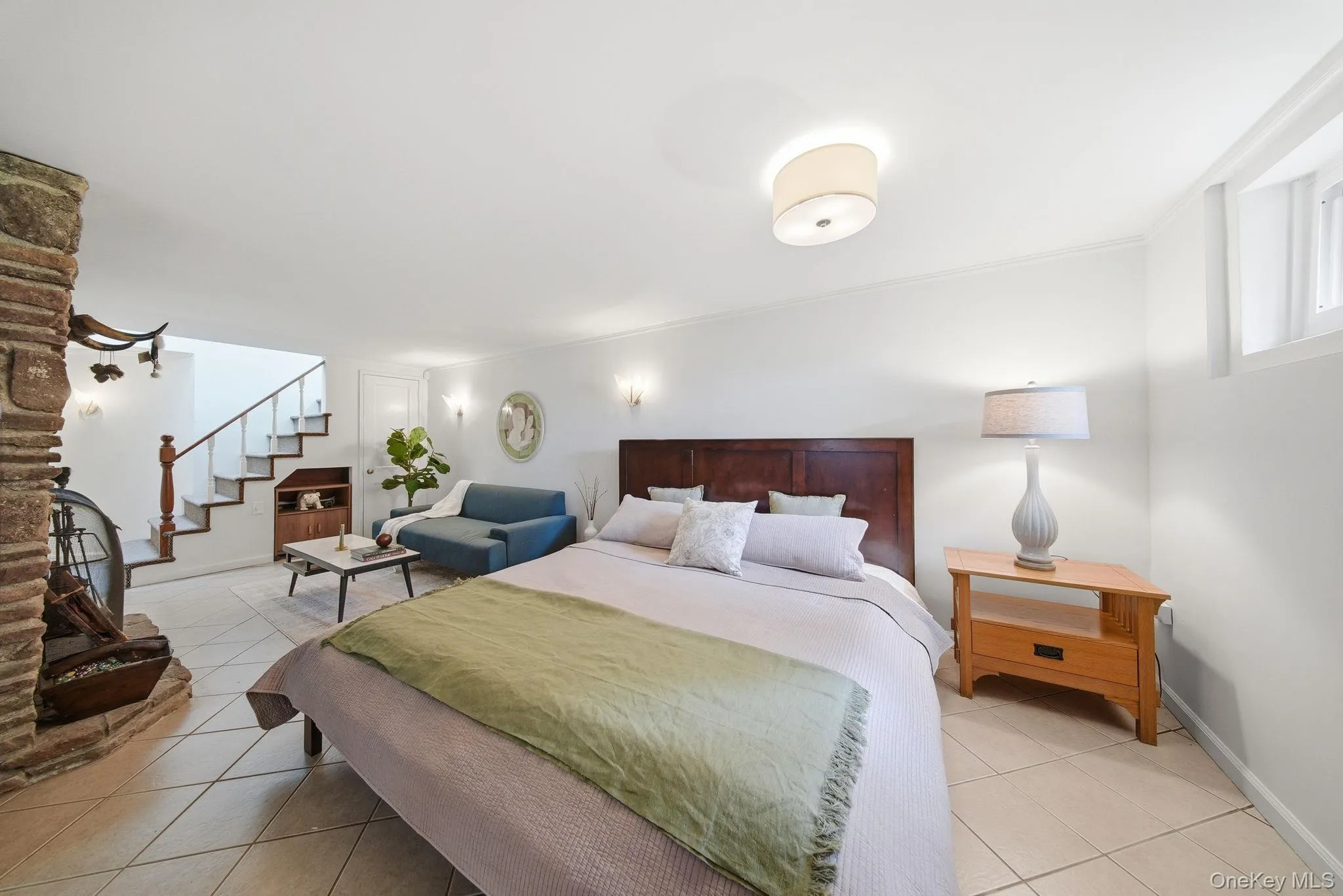 Spacious room featuring tile flooring, a stone fireplace, a wood-finish staircase with white risers, and recessed wall sconces Spacious room featuring tile flooring, a stone fireplace, a wood-finish staircase with white risers, and recessed wall sconces