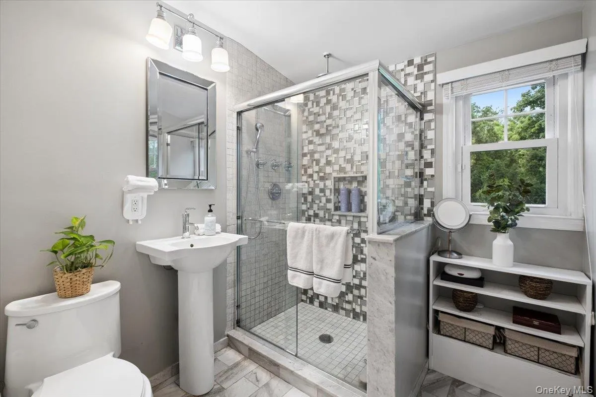 Glass-enclosed shower with mosaic tile surround and integrated niche, complemented by a pedestal sink, framed mirror, and track lighting. Glass-enclosed shower with mosaic tile surround and integrated niche, complemented by a pedestal sink, framed mirror, and track lighting.