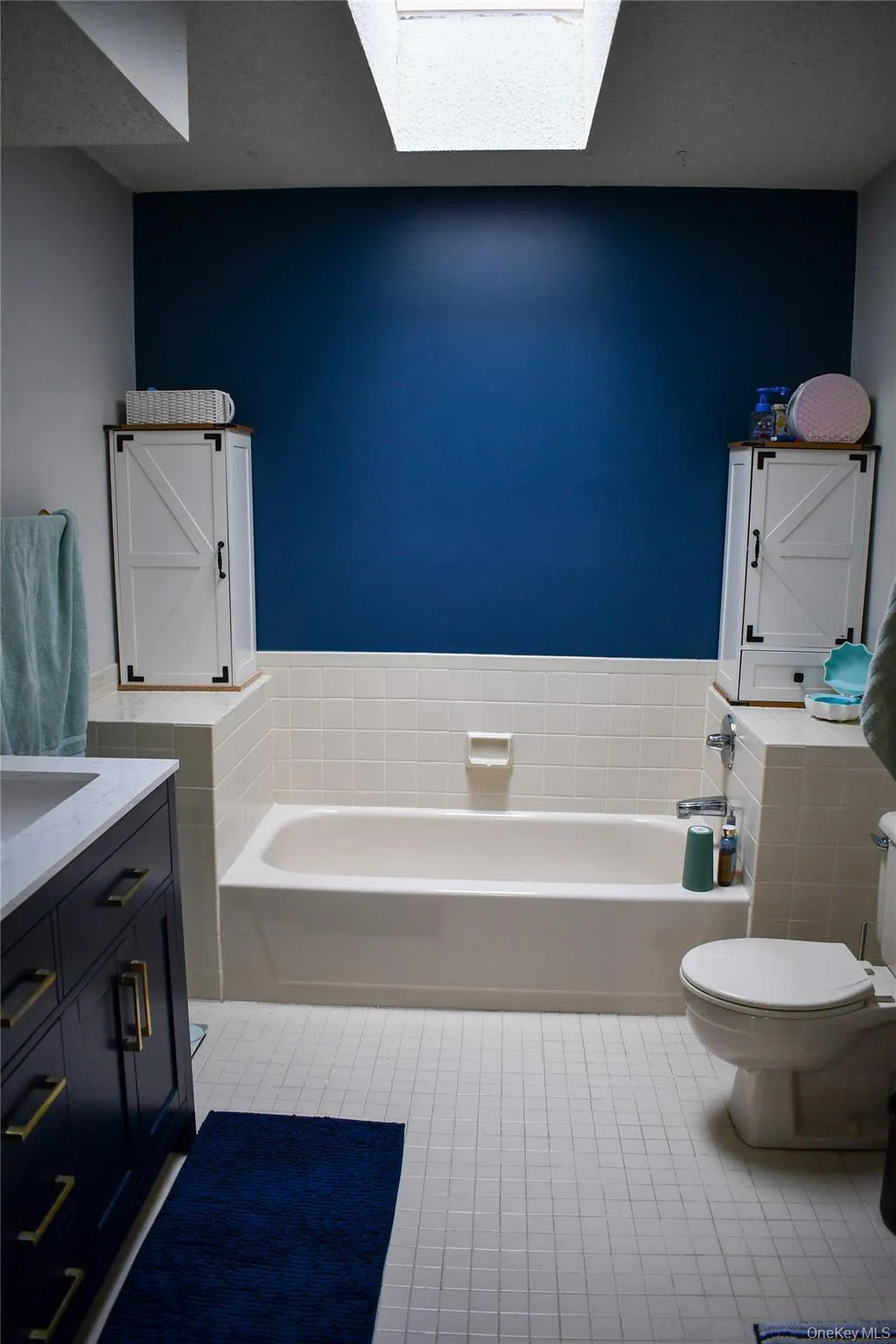 Bathroom featuring a built-in bathtub with white tile surround, white square tile flooring, a dark blue accent wall, and a skylight Bathroom featuring a built-in bathtub with white tile surround, white square tile flooring, a dark blue accent wall, and a skylight