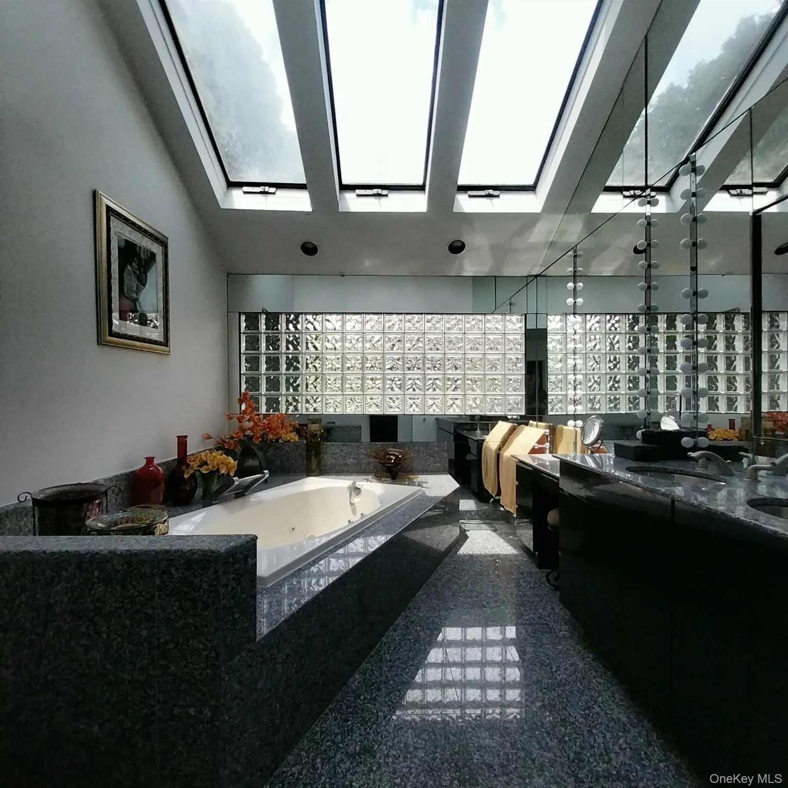 Bathroom featuring multiple skylights, a built-in tub with a stone surround, glass block wall, full-width mirror, and a dark stone tile floor Bathroom featuring multiple skylights, a built-in tub with a stone surround, glass block wall, full-width mirror, and a dark stone tile floor