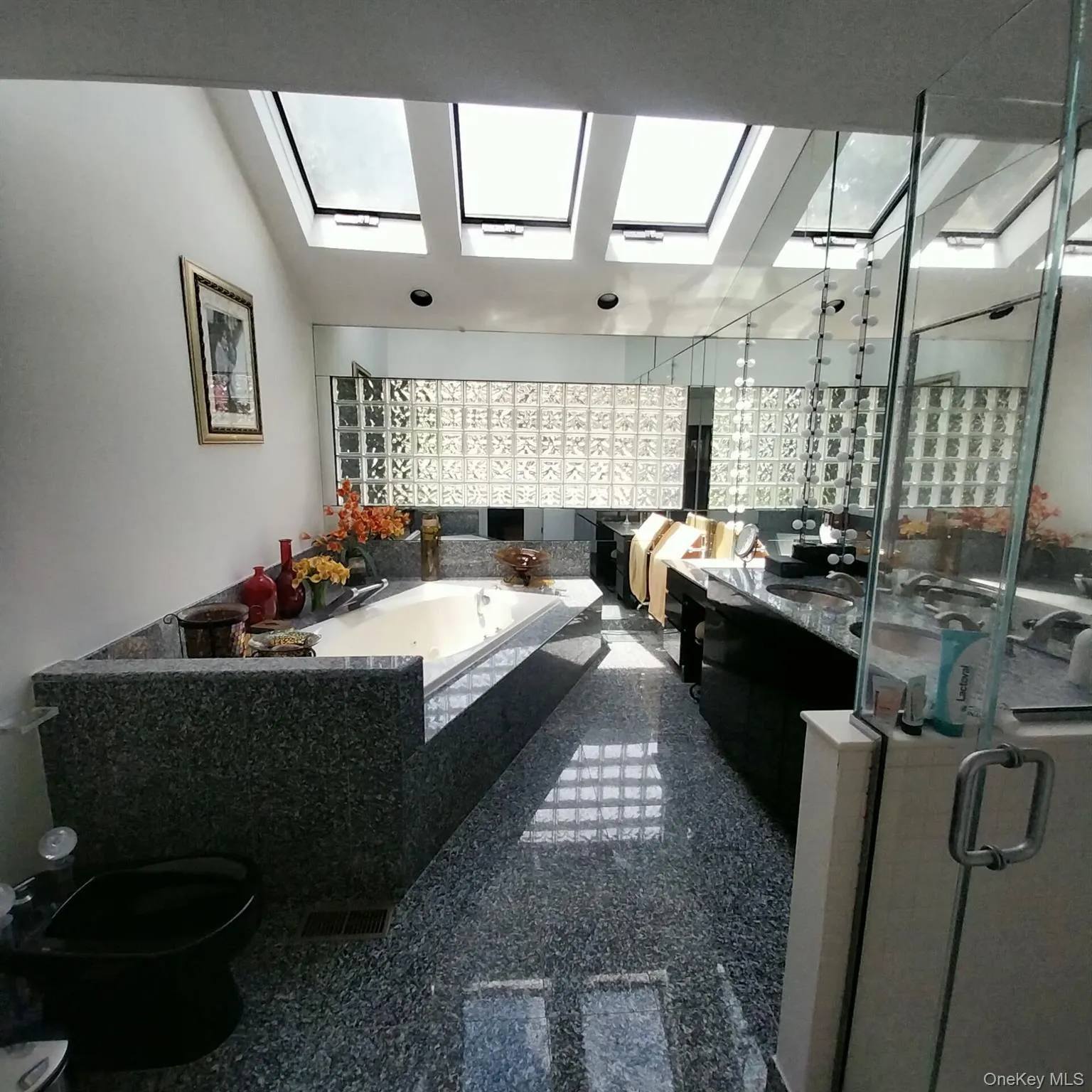 Bathroom featuring multiple skylights, a built-in tub with a stone surround, a glass block wall, and reflective flooring Bathroom featuring multiple skylights, a built-in tub with a stone surround, a glass block wall, and reflective flooring