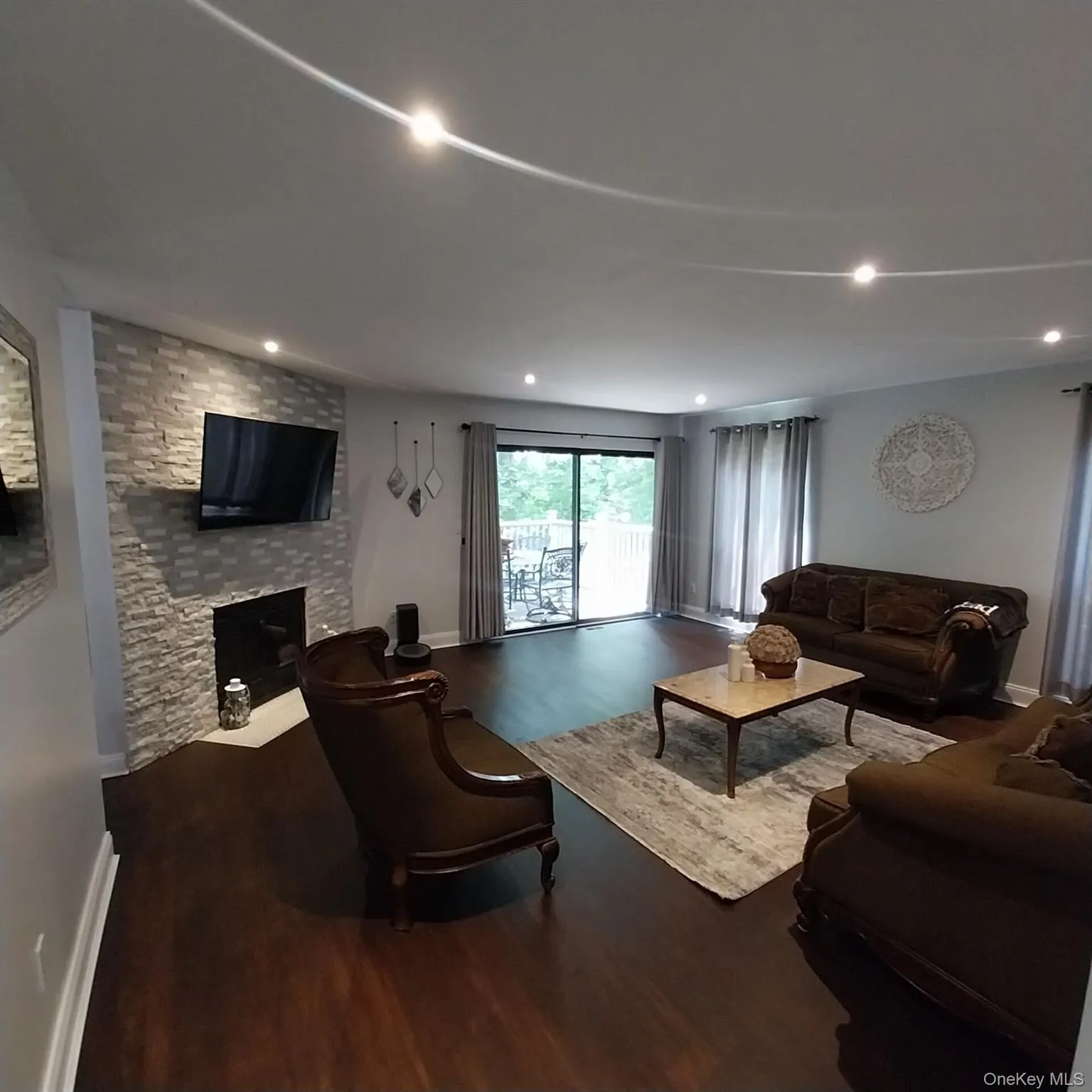 Spacious living area featuring wood-finish flooring, a stacked stone fireplace, recessed lighting, and sliding glass doors opening to an exterior deck Spacious living area featuring wood-finish flooring, a stacked stone fireplace, recessed lighting, and sliding glass doors opening to an exterior deck