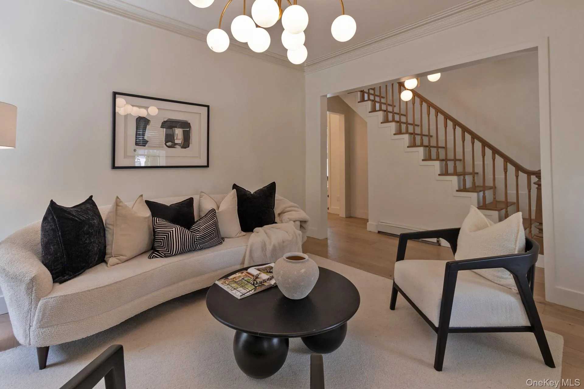 Open-concept living area featuring light wood-finish flooring, a contemporary chandelier with multiple globe lights, and an elegant staircase with wood newel posts and balusters Open-concept living area featuring light wood-finish flooring, a contemporary chandelier with multiple globe lights, and an elegant staircase with wood newel posts and balusters
