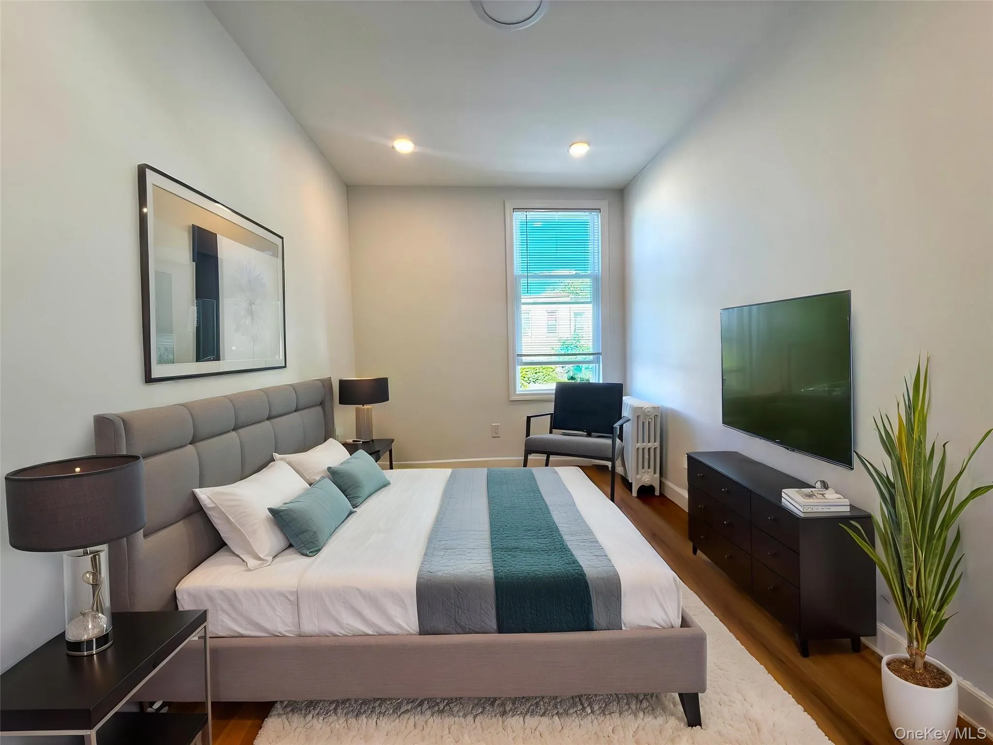 Bright bedroom featuring recessed lighting, a single window with blinds, wood-finish flooring, and light-toned walls Bright bedroom featuring recessed lighting, a single window with blinds, wood-finish flooring, and light-toned walls