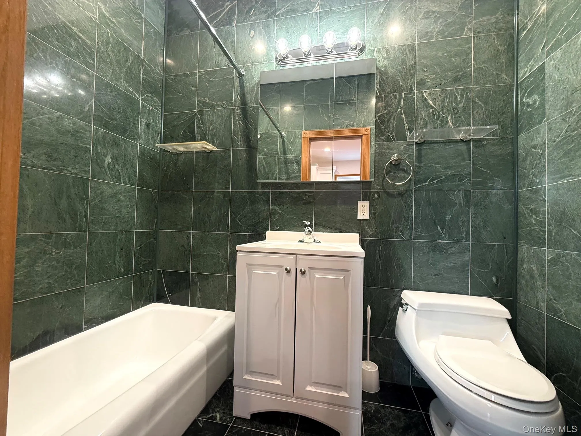 Bathroom featuring green stone tile walls, a white vanity with paneling, a white toilet, a white bathtub, and a mirrored medicine cabinet Bathroom featuring green stone tile walls, a white vanity with paneling, a white toilet, a white bathtub, and a mirrored medicine cabinet