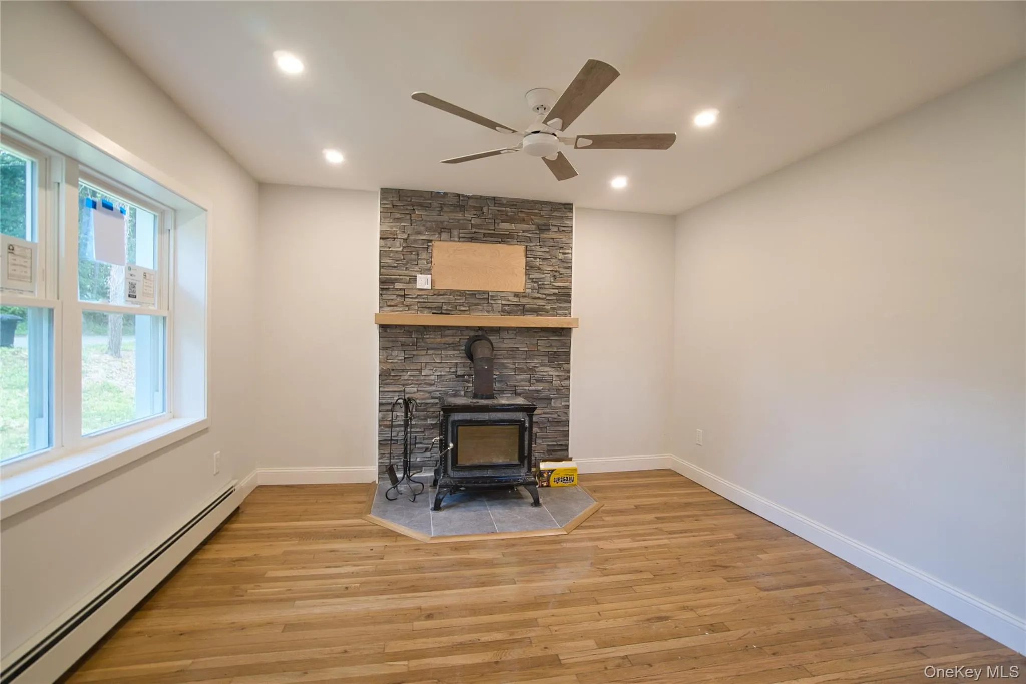 Living space featuring a stone-clad accent wall with wood stove, wood-finish flooring, a ceiling fan, recessed lighting, and white trim Living space featuring a stone-clad accent wall with wood stove, wood-finish flooring, a ceiling fan, recessed lighting, and white trim