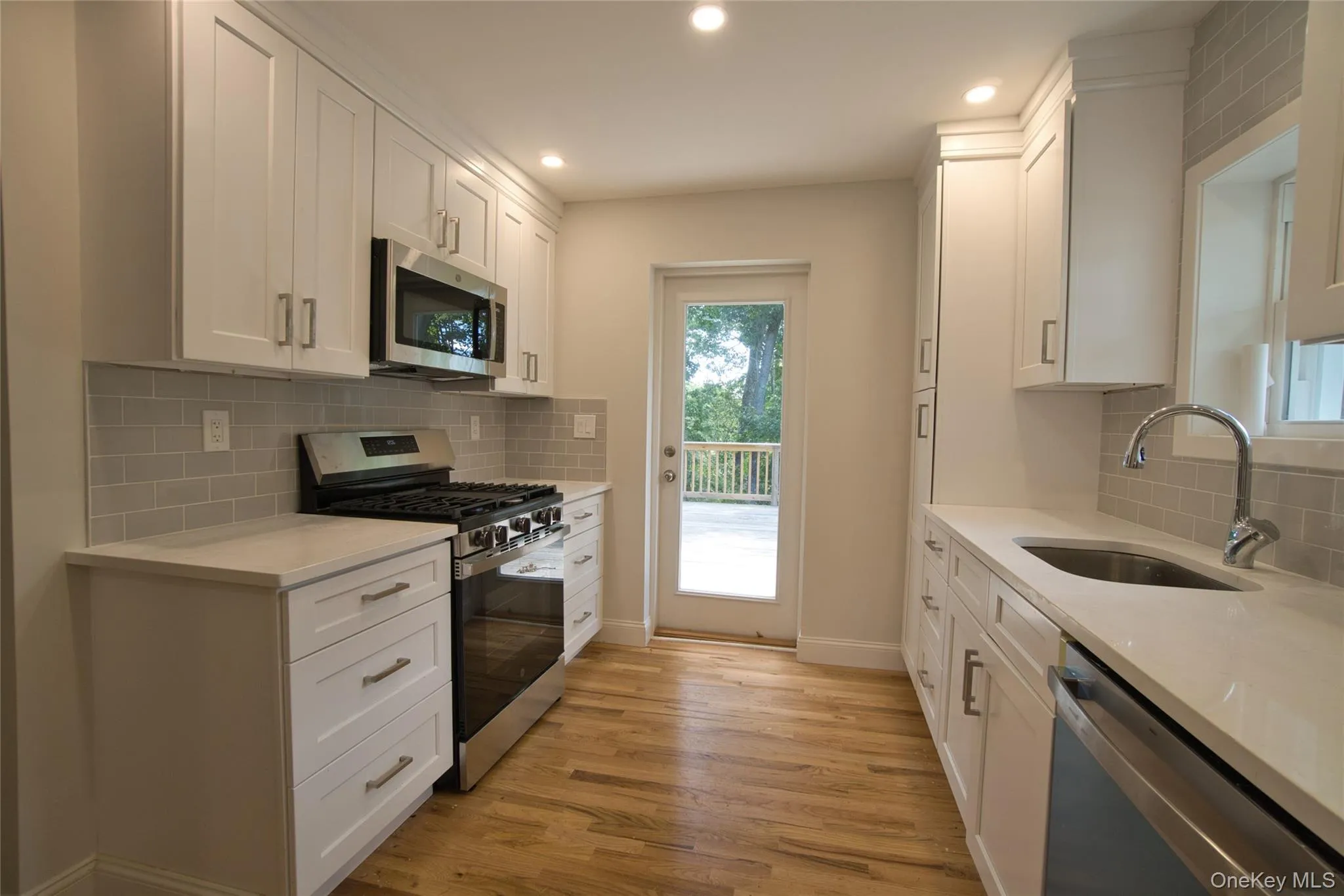 Galley kitchen featuring white shaker cabinetry, stainless steel appliances, a light gray subway tile backsplash, and light wood-finish flooring Galley kitchen featuring white shaker cabinetry, stainless steel appliances, a light gray subway tile backsplash, and light wood-finish flooring