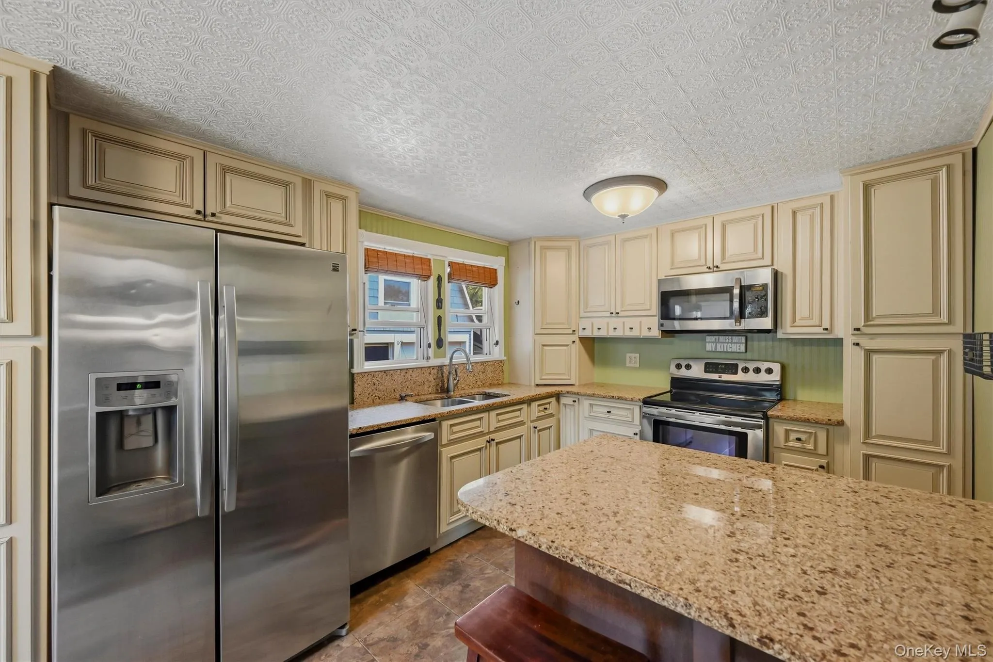 Kitchen featuring a granite-topped island, stainless steel appliances, light-toned cabinetry, a textured ceiling, and an integrated sink with a window backdrop Kitchen featuring a granite-topped island, stainless steel appliances, light-toned cabinetry, a textured ceiling, and an integrated sink with a window backdrop