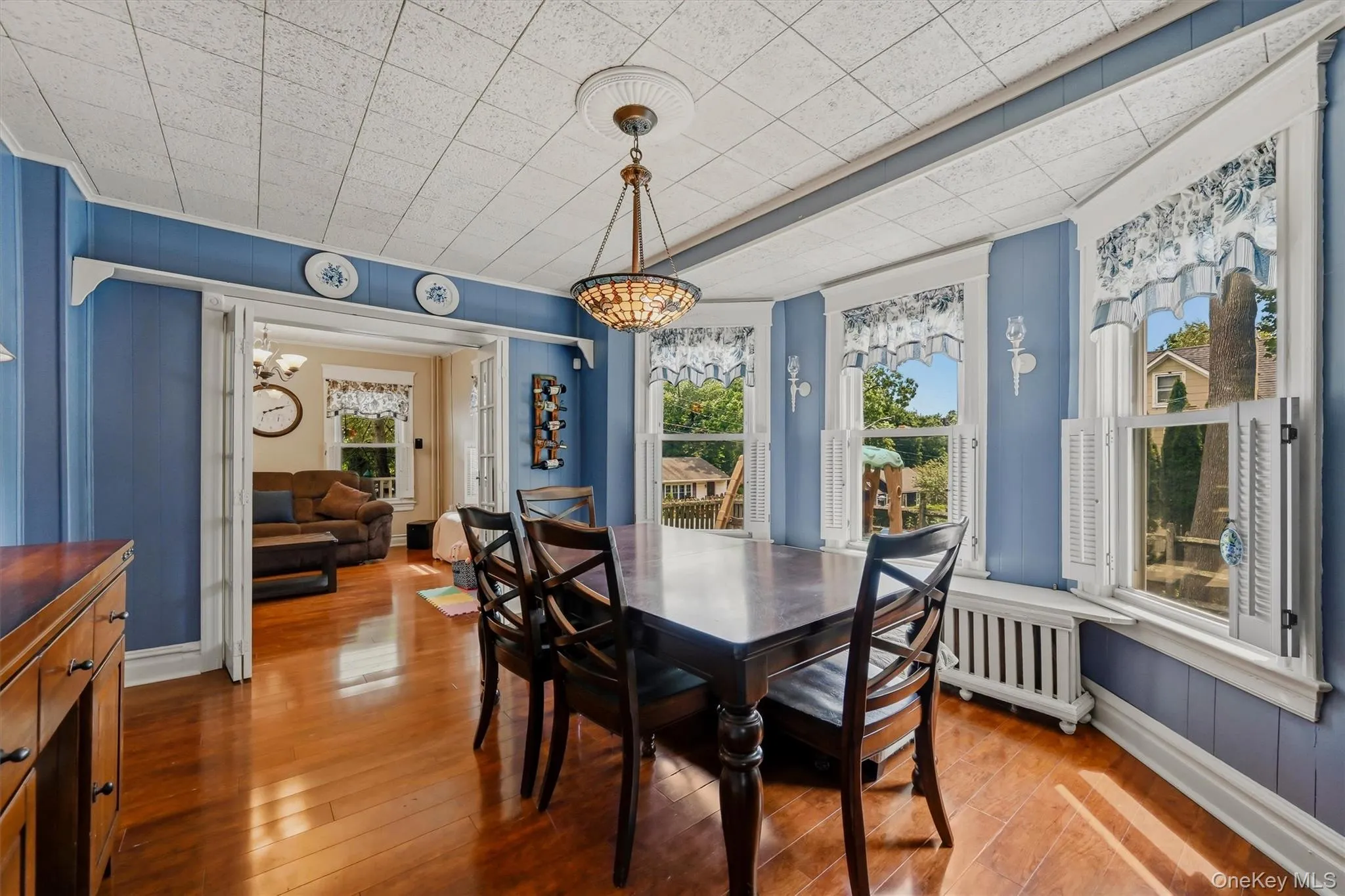 Dining area featuring hardwood flooring, a decorative ceiling with a central chandelier, and a triple bay window with interior shutters Dining area featuring hardwood flooring, a decorative ceiling with a central chandelier, and a triple bay window with interior shutters