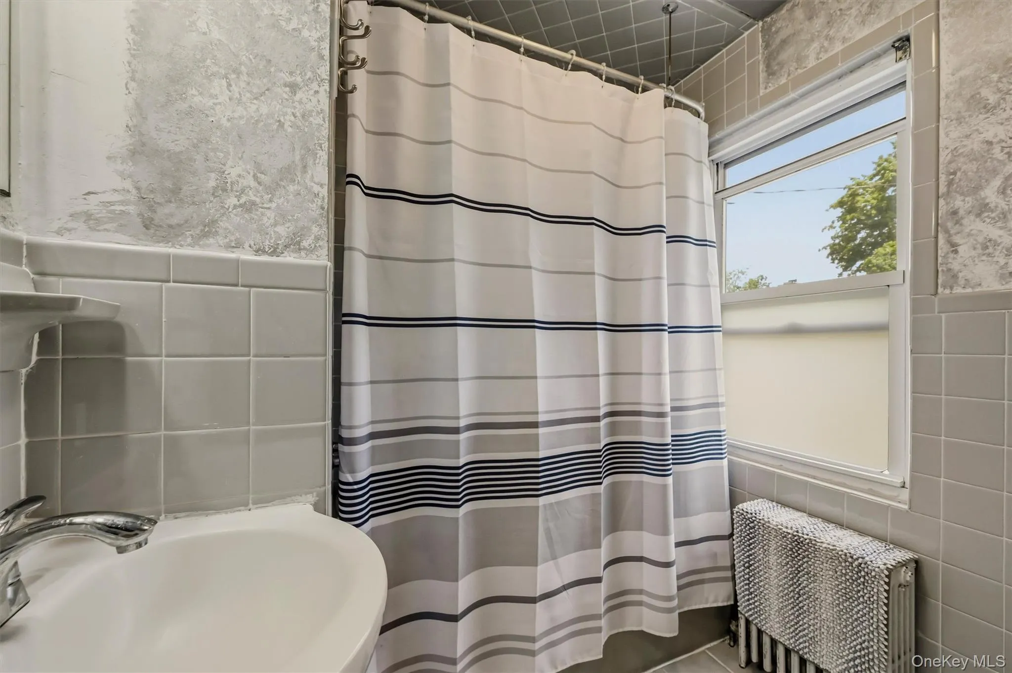 Bathroom featuring light grey wall tiling, a white basin sink with chrome fixtures, and a window providing natural illumination Bathroom featuring light grey wall tiling, a white basin sink with chrome fixtures, and a window providing natural illumination
