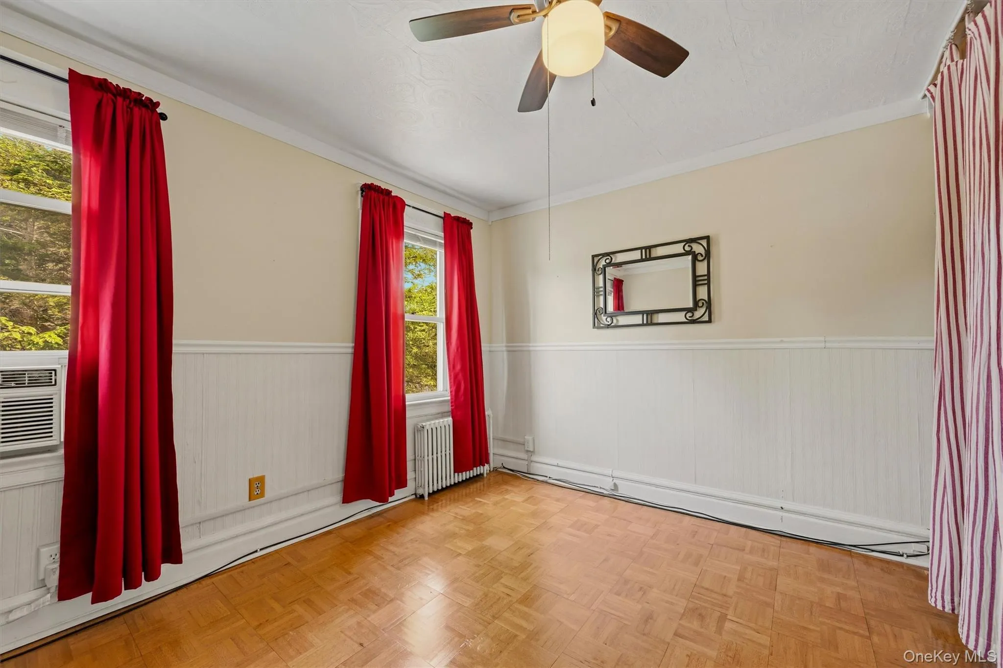 Room featuring a ceiling fan with integrated lighting, wood-finish parquet flooring, two windows with window treatments, and white wainscoting Room featuring a ceiling fan with integrated lighting, wood-finish parquet flooring, two windows with window treatments, and white wainscoting