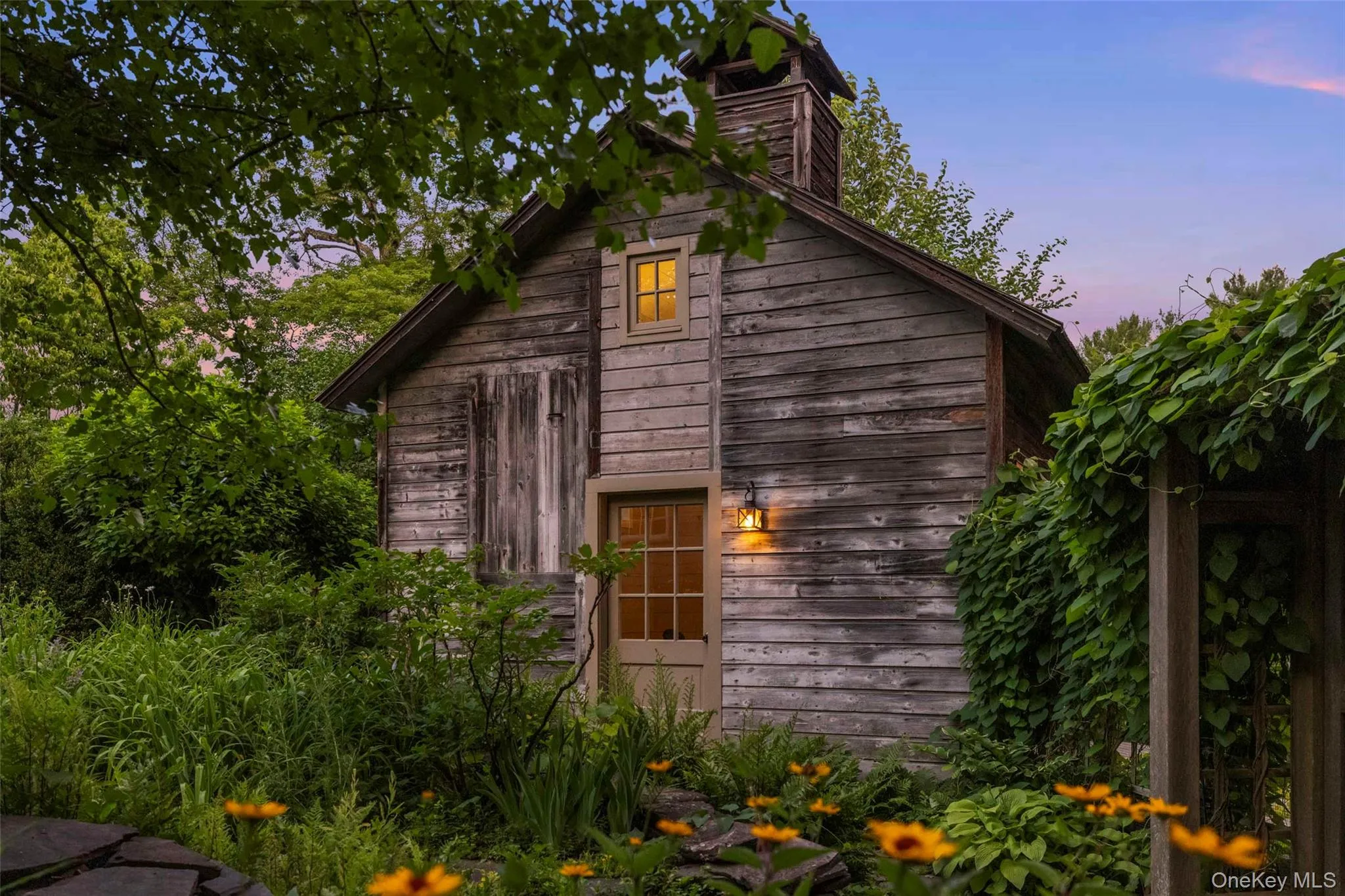 As evening falls, the wellness studio glows warmly amid mature gardens and lush landscaping As evening falls, the wellness studio glows warmly amid mature gardens and lush landscaping