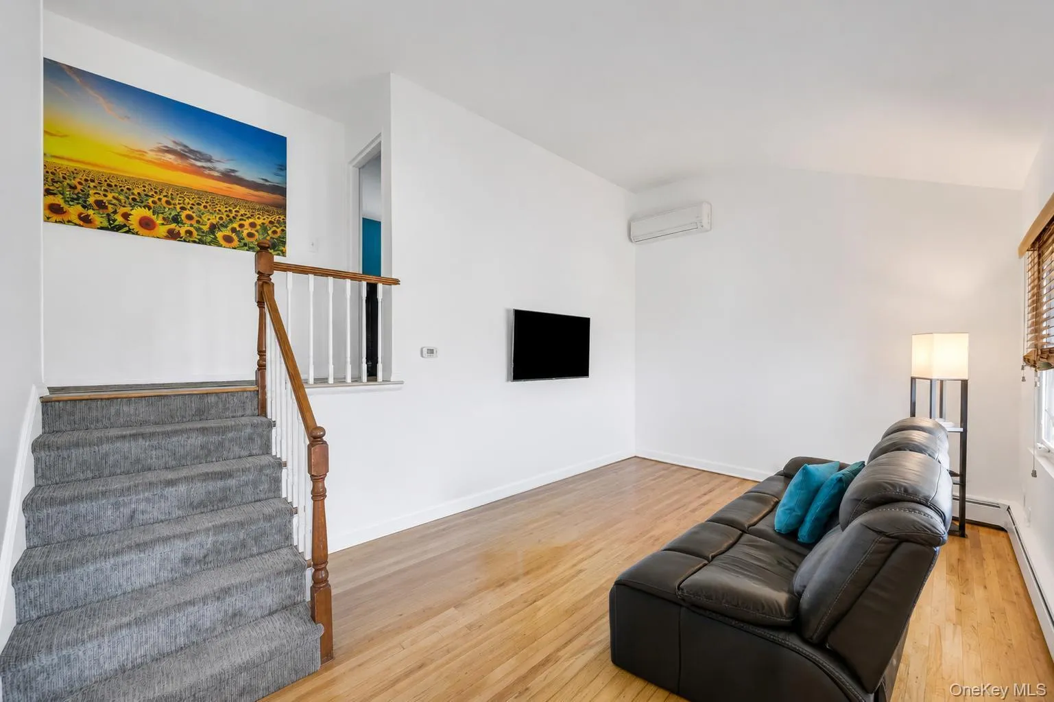 Living area featuring wood-finish flooring, a wall-mounted air conditioning unit, and a staircase with a wood handrail and carpeted treads Living area featuring wood-finish flooring, a wall-mounted air conditioning unit, and a staircase with a wood handrail and carpeted treads