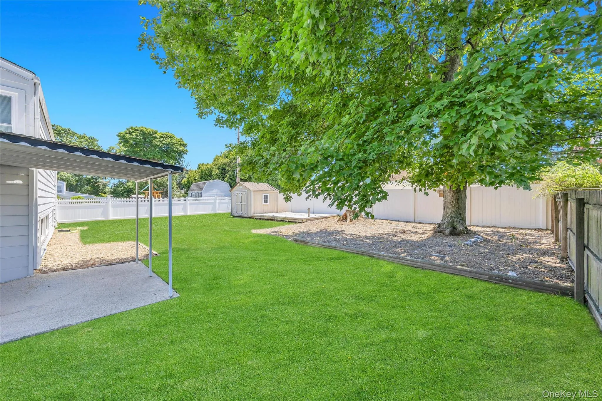 Expansive backyard featuring a manicured lawn, a large mature tree, a white privacy fence, a covered patio area, and a storage shed on a raised deck Expansive backyard featuring a manicured lawn, a large mature tree, a white privacy fence, a covered patio area, and a storage shed on a raised deck