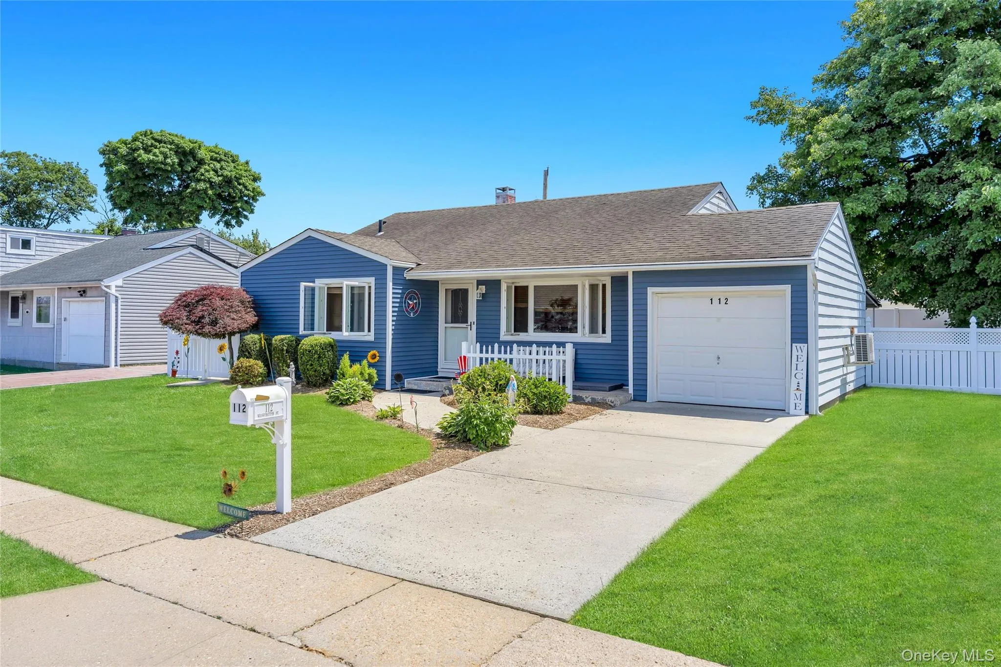 Blue-sided residence featuring a dark shingle roof, attached garage, white picket fence, and a concrete driveway Blue-sided residence featuring a dark shingle roof, attached garage, white picket fence, and a concrete driveway