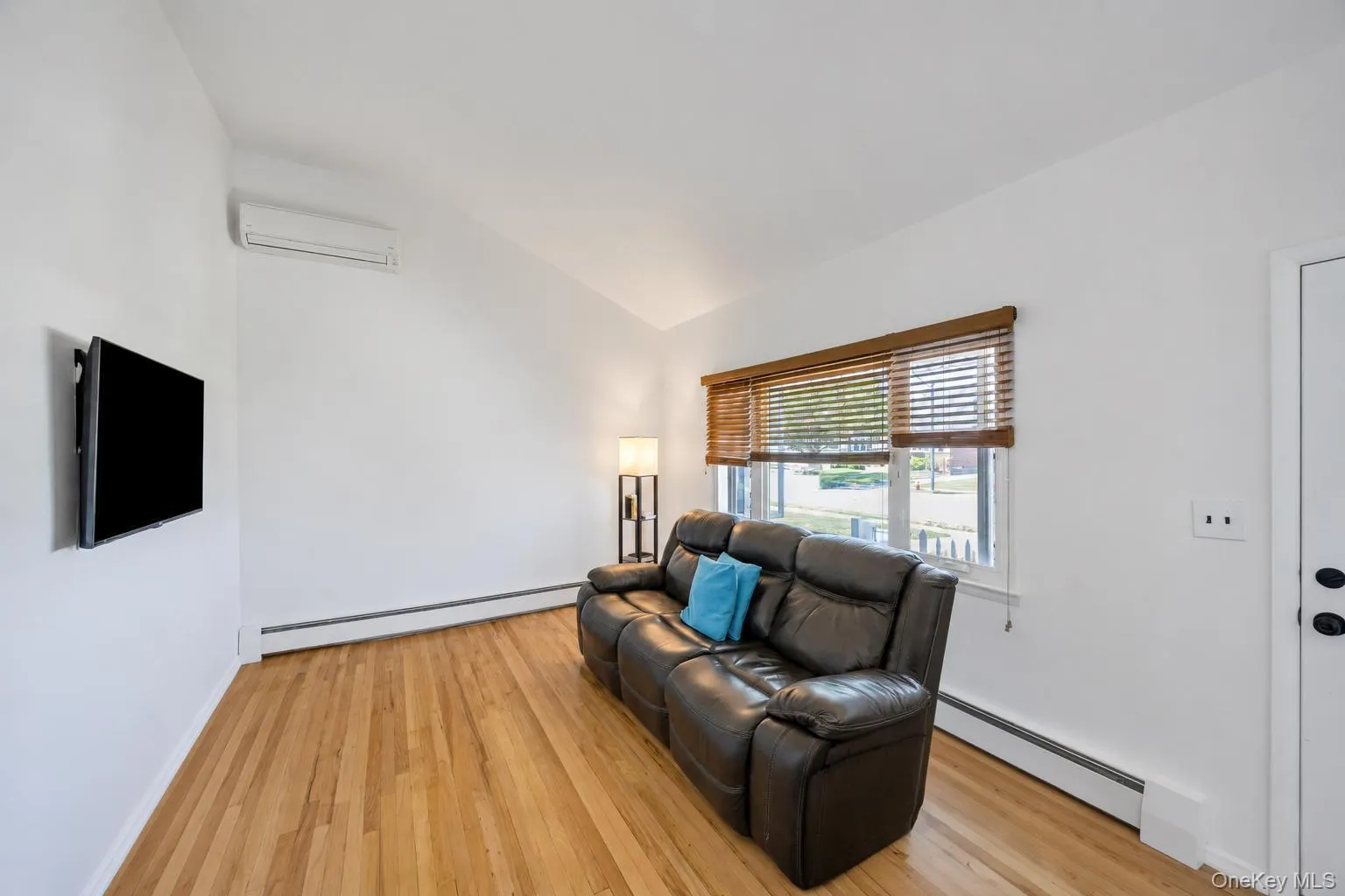 Bright interior space featuring wood-finish flooring, a wall-mounted climate control unit, and a window with wood-slat blinds Bright interior space featuring wood-finish flooring, a wall-mounted climate control unit, and a window with wood-slat blinds