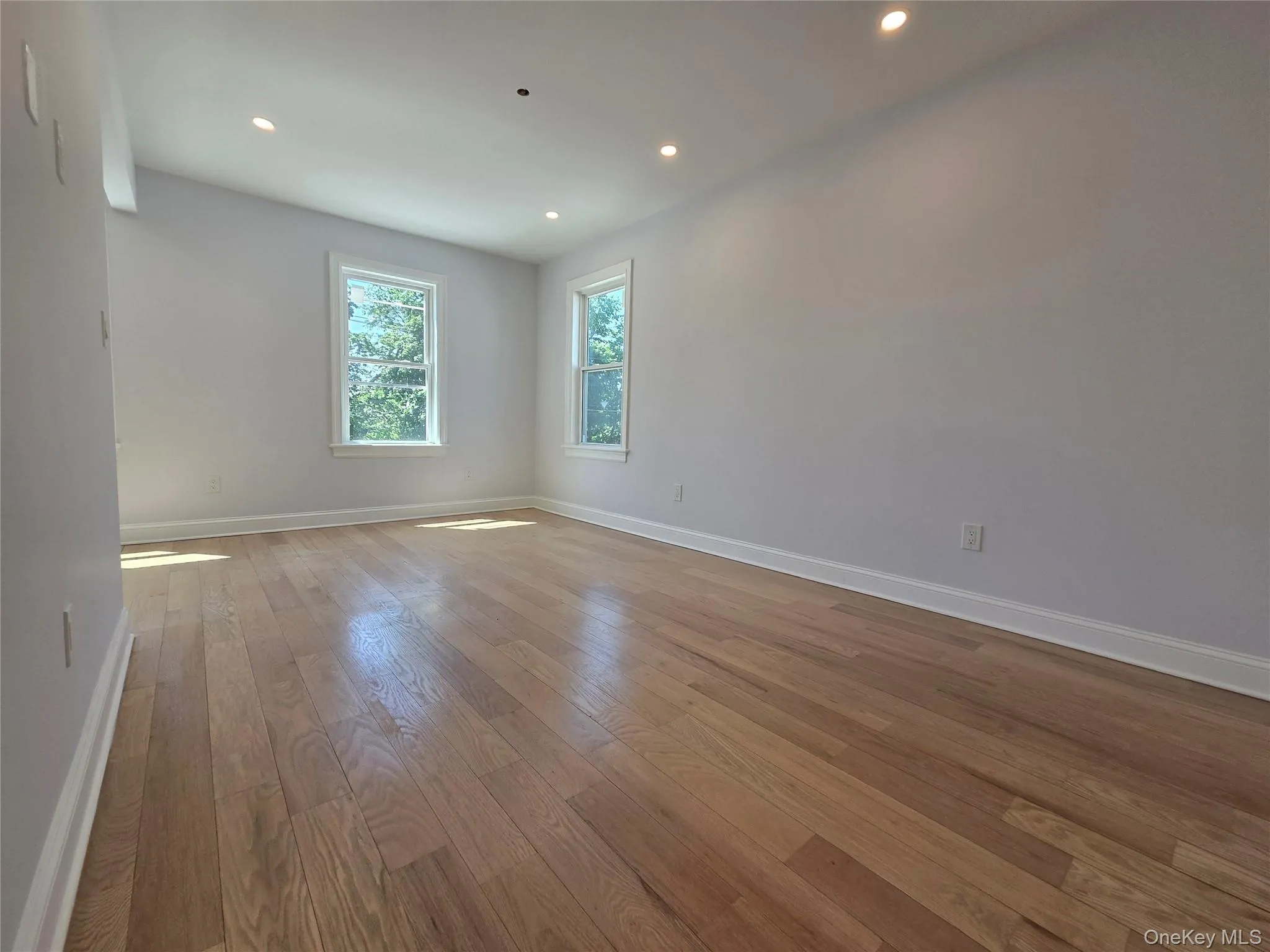 Light-filled room featuring wood-finish flooring, two windows with white trim, recessed ceiling lighting, and crisp white baseboards Light-filled room featuring wood-finish flooring, two windows with white trim, recessed ceiling lighting, and crisp white baseboards