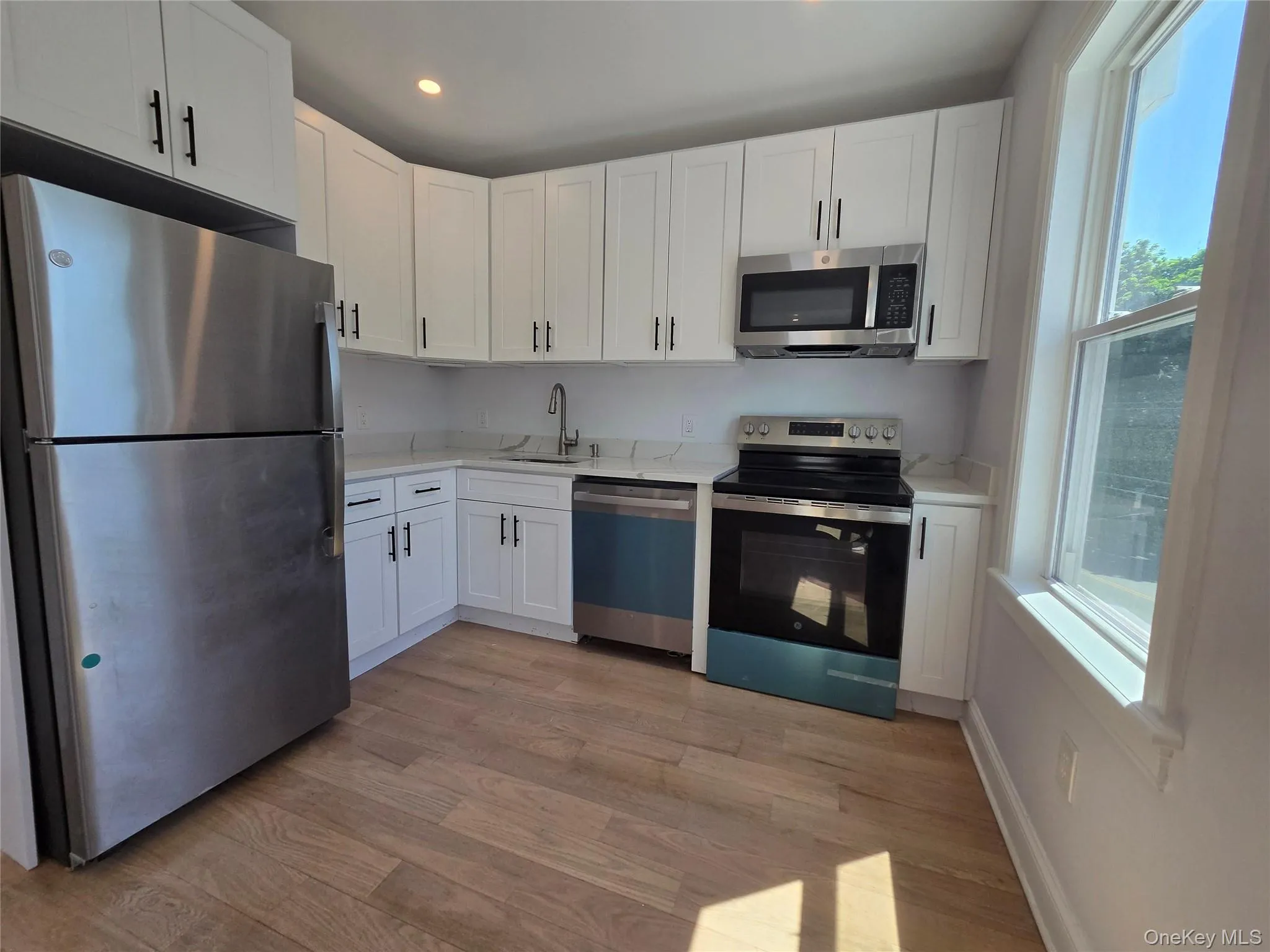 Kitchen featuring white cabinetry with dark hardware, a stainless steel refrigerator, stainless steel microwave, and wood-finish flooring Kitchen featuring white cabinetry with dark hardware, a stainless steel refrigerator, stainless steel microwave, and wood-finish flooring