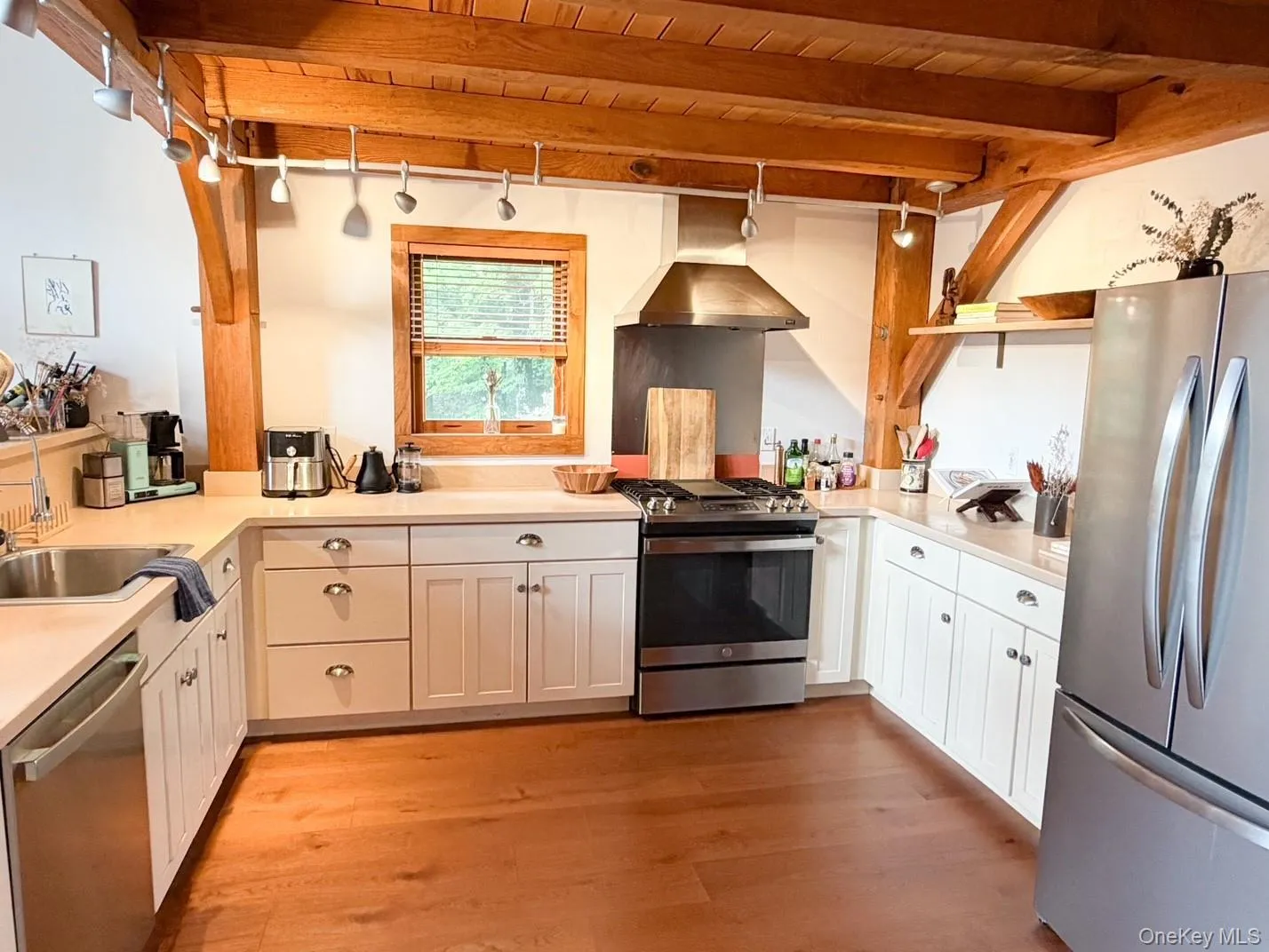 Kitchen featuring wood-finish flooring, exposed wood beams, white cabinetry with silver hardware, stainless steel appliances, and a window with wood trim Kitchen featuring wood-finish flooring, exposed wood beams, white cabinetry with silver hardware, stainless steel appliances, and a window with wood trim