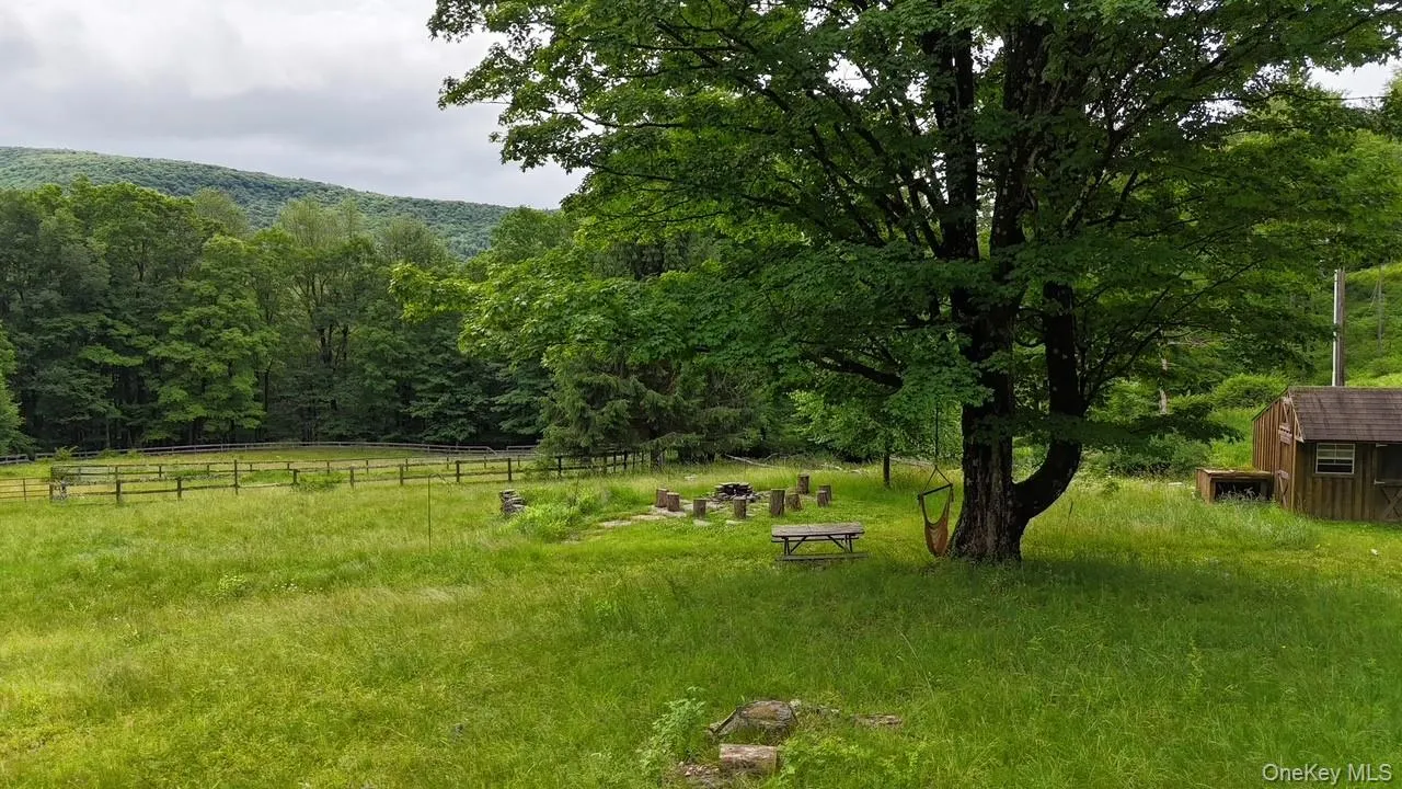 Expansive green field featuring a large mature tree, a rustic wooden outbuilding, and distant forested hills Expansive green field featuring a large mature tree, a rustic wooden outbuilding, and distant forested hills