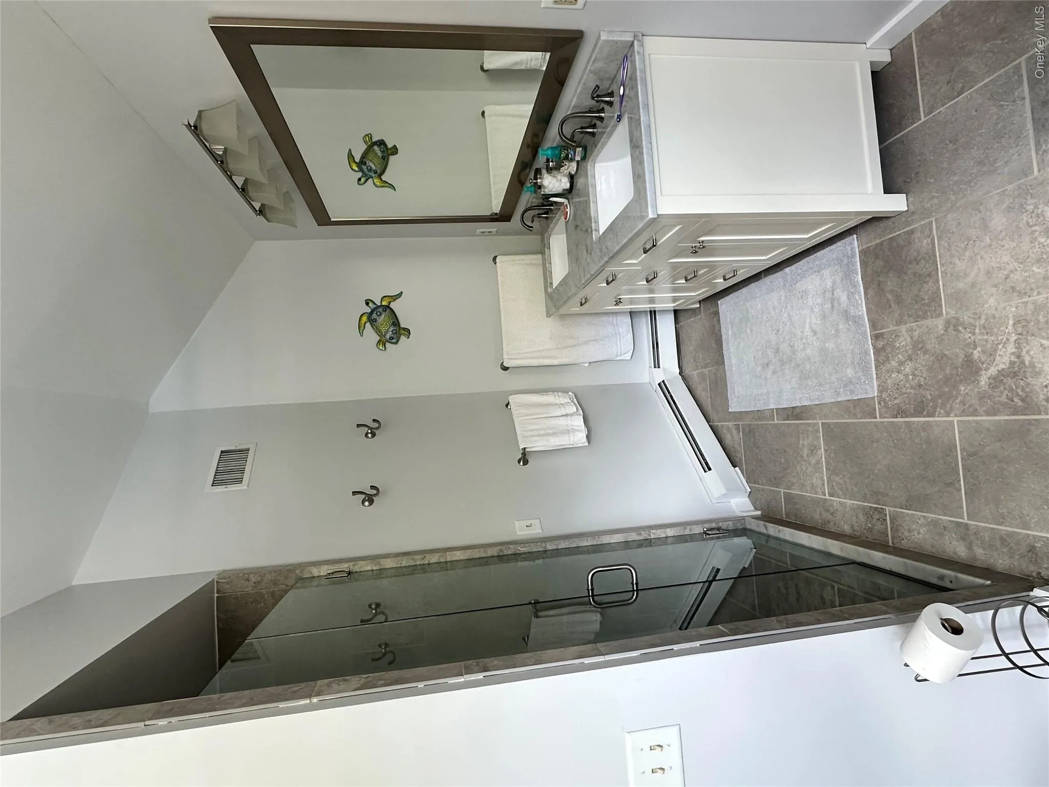 Bathroom featuring a large glass-enclosed shower with tile surround, a double vanity with undermount sinks and a stone countertop, a framed mirror, and large format floor tiling Bathroom featuring a large glass-enclosed shower with tile surround, a double vanity with undermount sinks and a stone countertop, a framed mirror, and large format floor tiling