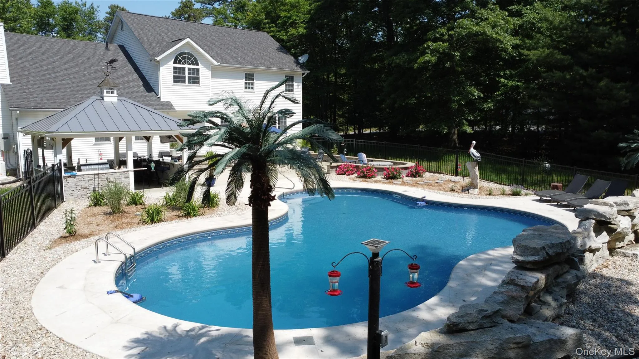 Freeform salt water swimming pool featuring a white concrete surround, adjacent stone retaining wall, and a nearby covered patio structure with a dark roof Freeform salt water swimming pool featuring a white concrete surround, adjacent stone retaining wall, and a nearby covered patio structure with a dark roof