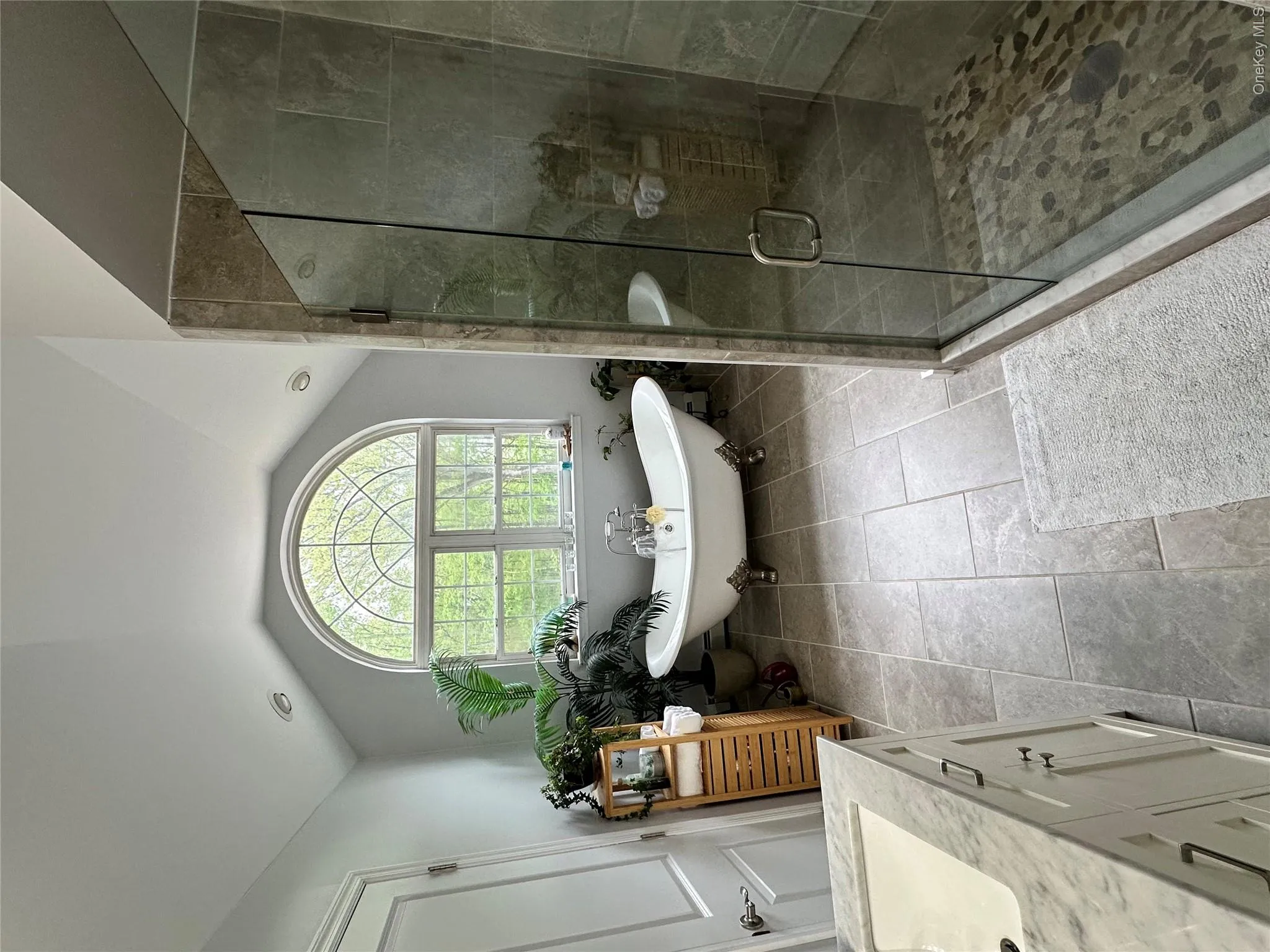 Spacious bathroom featuring a freestanding clawfoot tub, an arched window, a glass-enclosed shower with pebble flooring, a vanity with an integrated sink and stone countertop, and large format floor tiling Spacious bathroom featuring a freestanding clawfoot tub, an arched window, a glass-enclosed shower with pebble flooring, a vanity with an integrated sink and stone countertop, and large format floor tiling