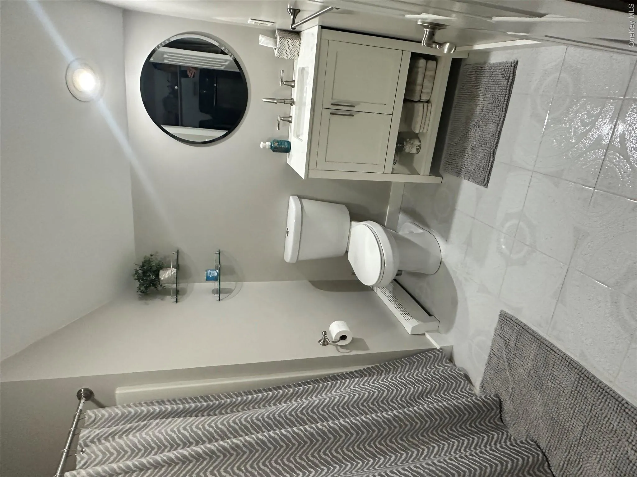 Full Bathroom featuring light gray tiled flooring, a white vanity with a rectangular sink and brushed nickel faucet, a round wall-mounted mirror, and glass corner shelving Full Bathroom featuring light gray tiled flooring, a white vanity with a rectangular sink and brushed nickel faucet, a round wall-mounted mirror, and glass corner shelving