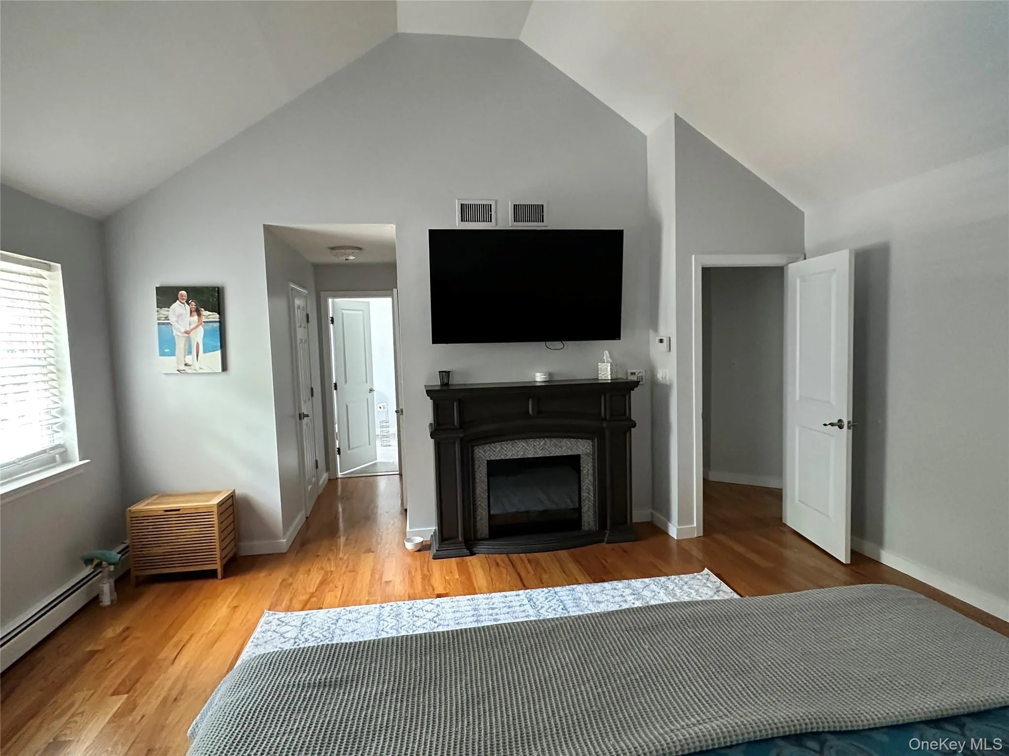 Room featuring vaulted ceilings, wood-finish flooring, a decorative electric fireplace with a dark wood-tone mantel, white interior doors, and a window with blinds Room featuring vaulted ceilings, wood-finish flooring, a decorative electric fireplace with a dark wood-tone mantel, white interior doors, and a window with blinds