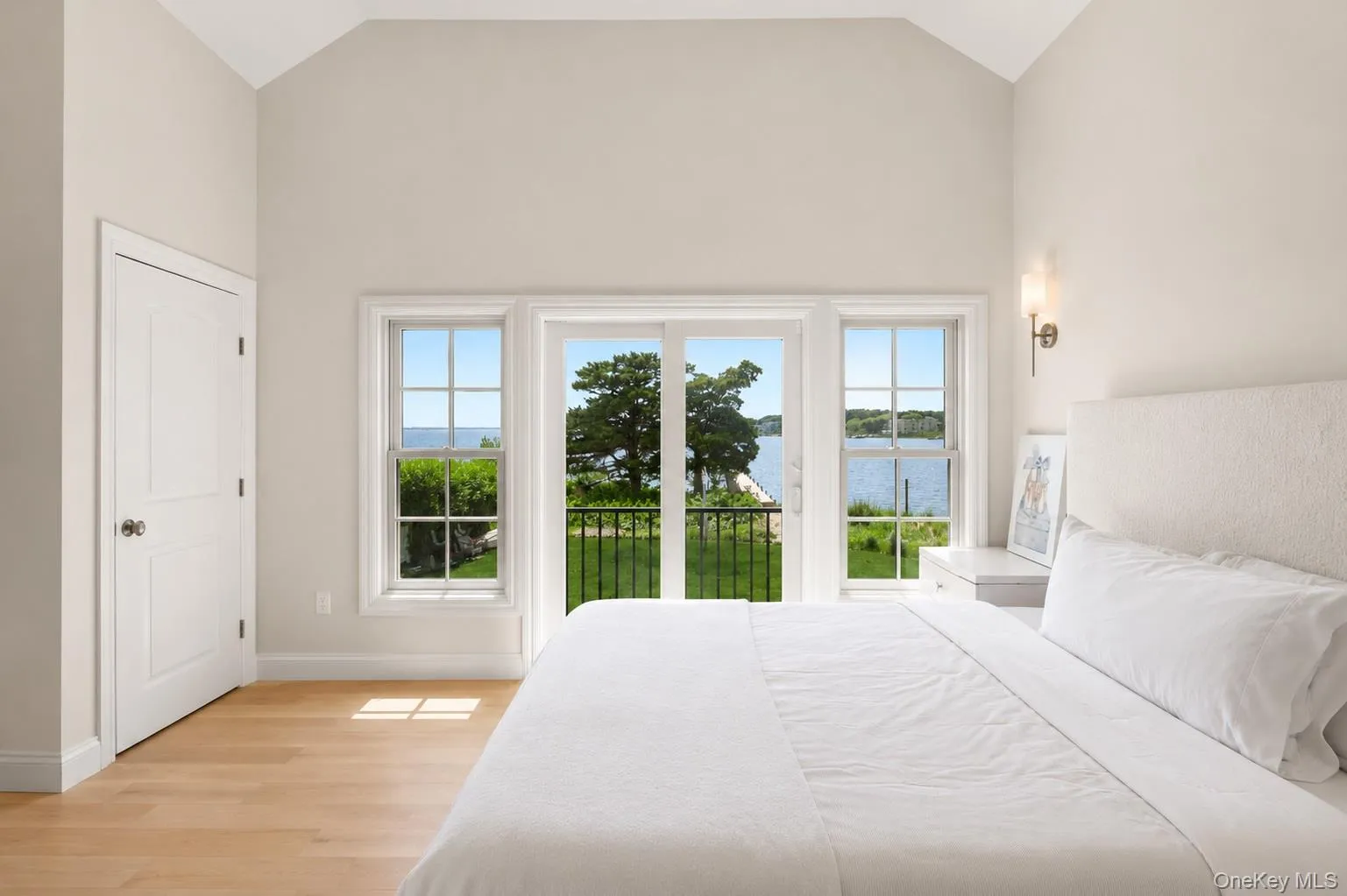 Vaulted ceiling bedroom featuring wood-finish flooring, a wall sconce, and a balcony with wrought iron railing overlooking a body of water Vaulted ceiling bedroom featuring wood-finish flooring, a wall sconce, and a balcony with wrought iron railing overlooking a body of water