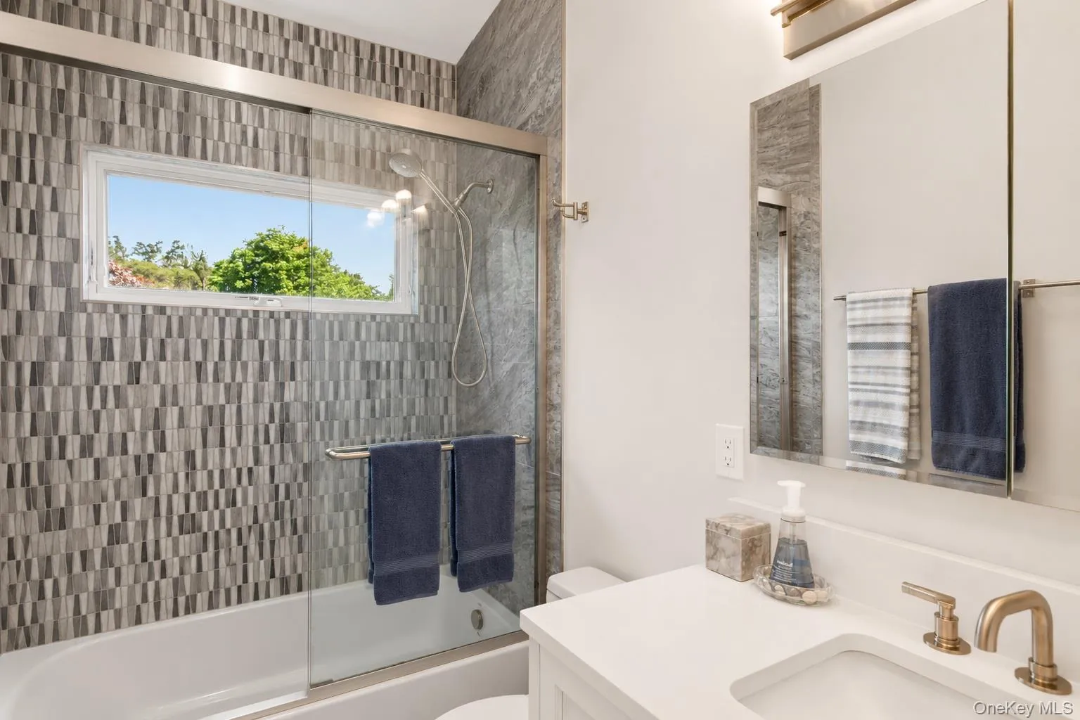 Contemporary bathroom featuring a combination shower and tub with sliding glass enclosure, mosaic-patterned tile surround, a horizontal window, and a single vanity with a white countertop and gold-toned fixtures Contemporary bathroom featuring a combination shower and tub with sliding glass enclosure, mosaic-patterned tile surround, a horizontal window, and a single vanity with a white countertop and gold-toned fixtures