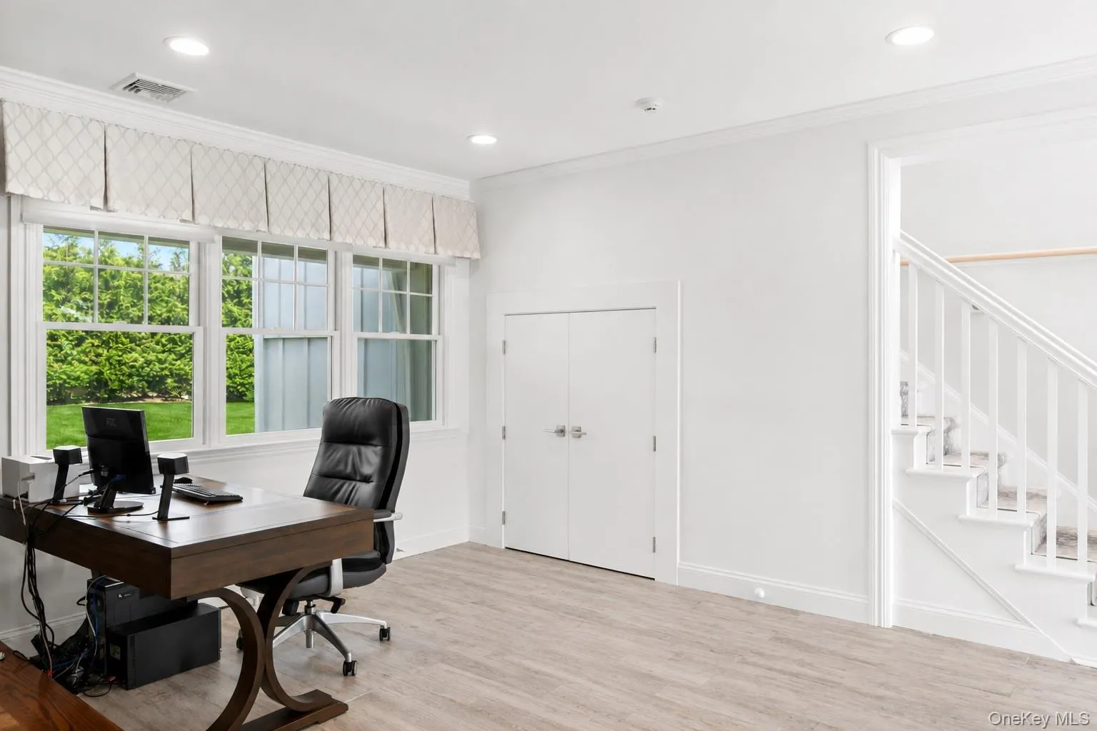 Bright room with light wood-finish flooring, featuring a large triple window with white trim, recessed lighting, and a white staircase with balusters Bright room with light wood-finish flooring, featuring a large triple window with white trim, recessed lighting, and a white staircase with balusters