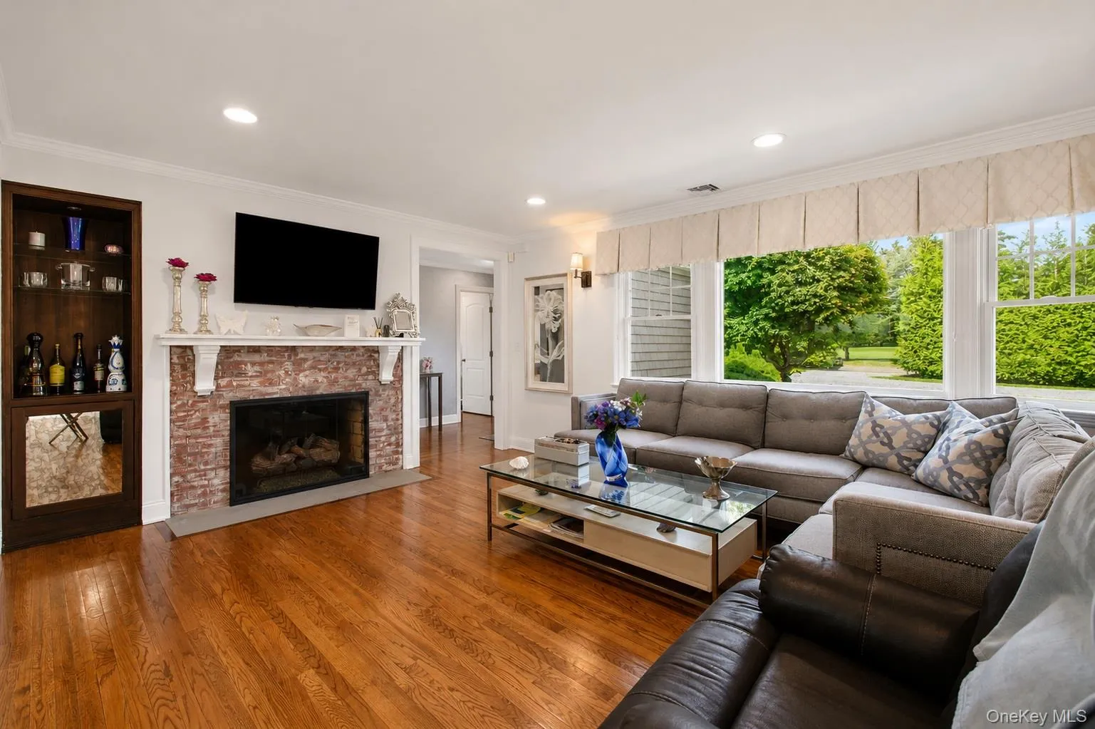 Living area featuring a red brick fireplace with a white mantel, recessed lighting, wood-finish flooring, a built-in display cabinet, and a large window arrangement Living area featuring a red brick fireplace with a white mantel, recessed lighting, wood-finish flooring, a built-in display cabinet, and a large window arrangement