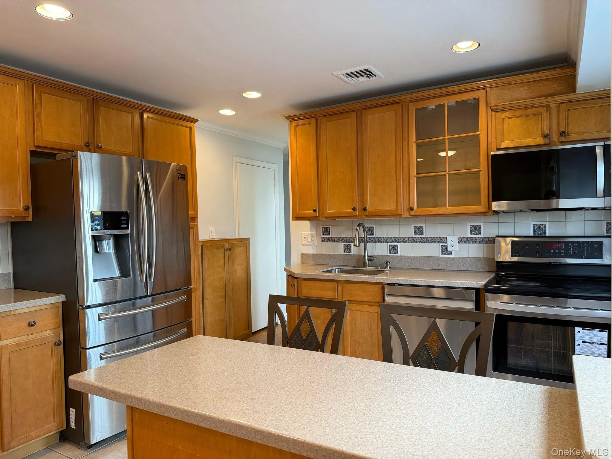 Kitchen with tasteful backsplash, stainless steel appliances, a sink, visible vents, and brown cabinetry Kitchen with tasteful backsplash, stainless steel appliances, a sink, visible vents, and brown cabinetry