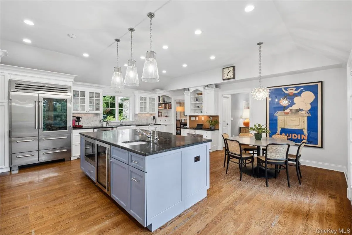 Gourmet kitchen featuring a large central island with integrated beverage refrigerator, stainless steel professional-grade refrigerator, wood-finish flooring, white cabinetry, and recessed lighting with easy access to the family room and outdoor space Gourmet kitchen featuring a large central island with integrated beverage refrigerator, stainless steel professional-grade refrigerator, wood-finish flooring, white cabinetry, and recessed lighting with easy access to the family room and outdoor space
