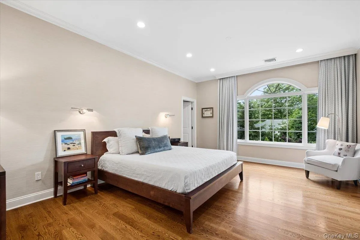 Light filled primary bedroom featuring a large arched window, polished wood-finish flooring, recessed lighting, crown molding, wall sconces and a large walk in closet Light filled primary bedroom featuring a large arched window, polished wood-finish flooring, recessed lighting, crown molding, wall sconces and a large walk in closet