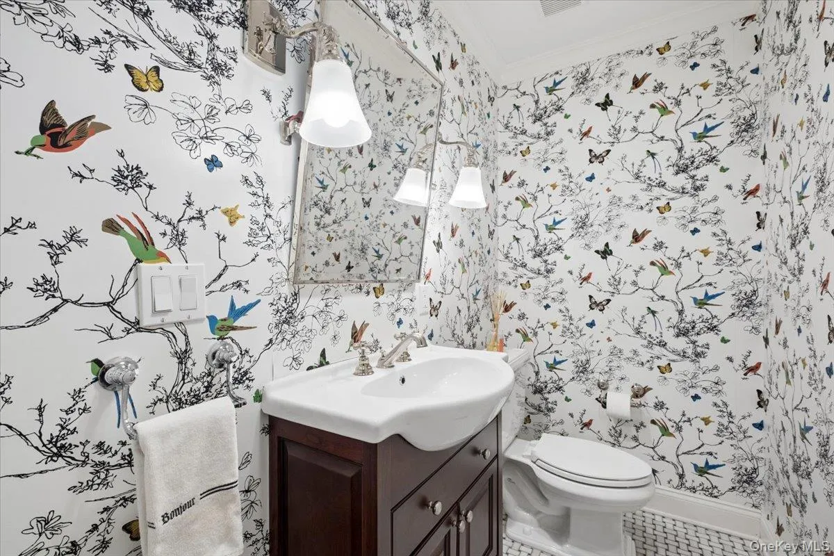Powder room featuring whimsical botanical-patterned wallpaper, a dark wood-finish vanity with an pedestal white sink, a wall-mounted mirror, brushed nickel sconces, and white hexagon tile flooring Powder room featuring whimsical botanical-patterned wallpaper, a dark wood-finish vanity with an pedestal white sink, a wall-mounted mirror, brushed nickel sconces, and white hexagon tile flooring